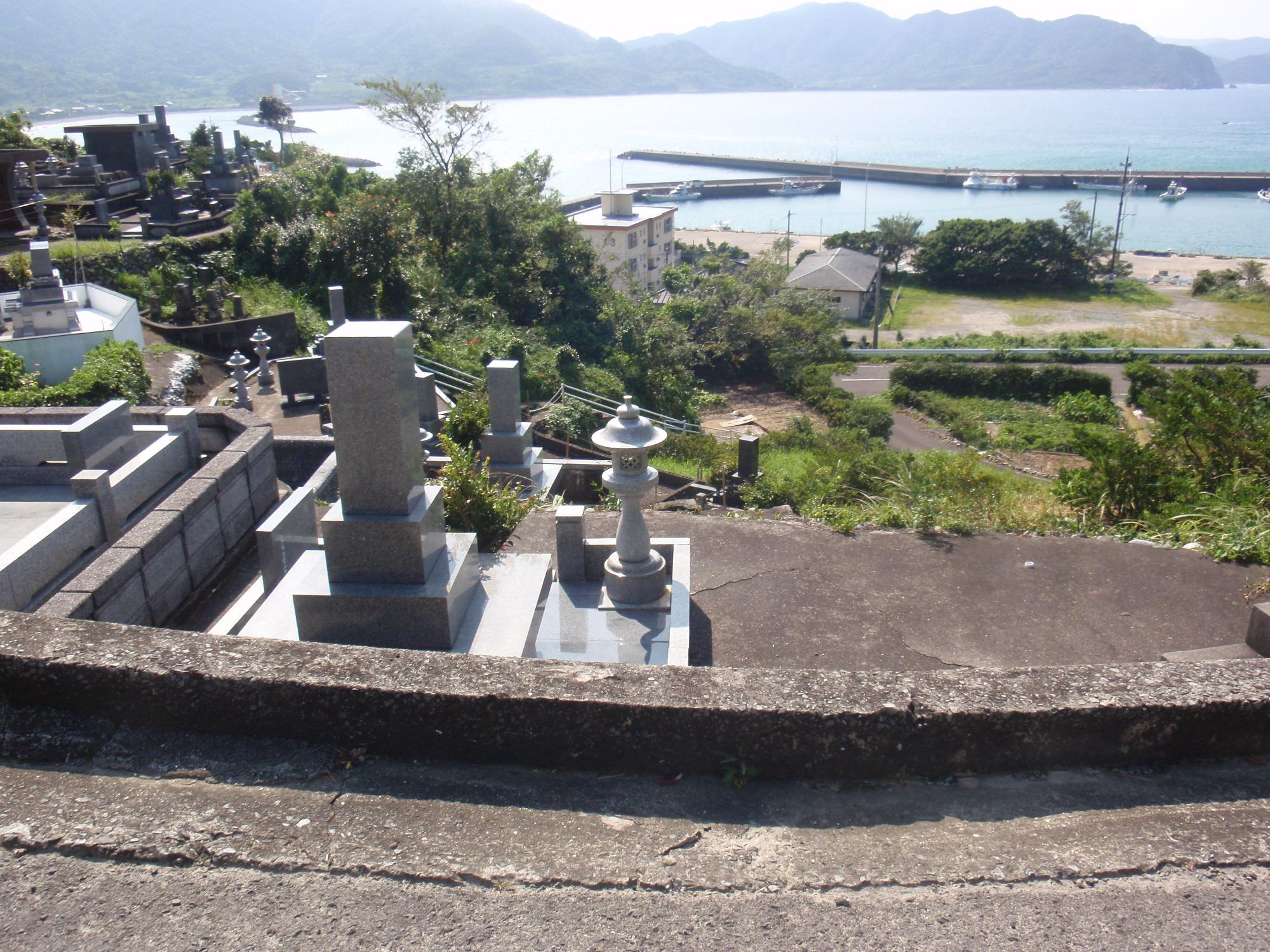 斜面に建つ海を見下ろすお墓の先に、海を囲うように山が続いている景色の写真