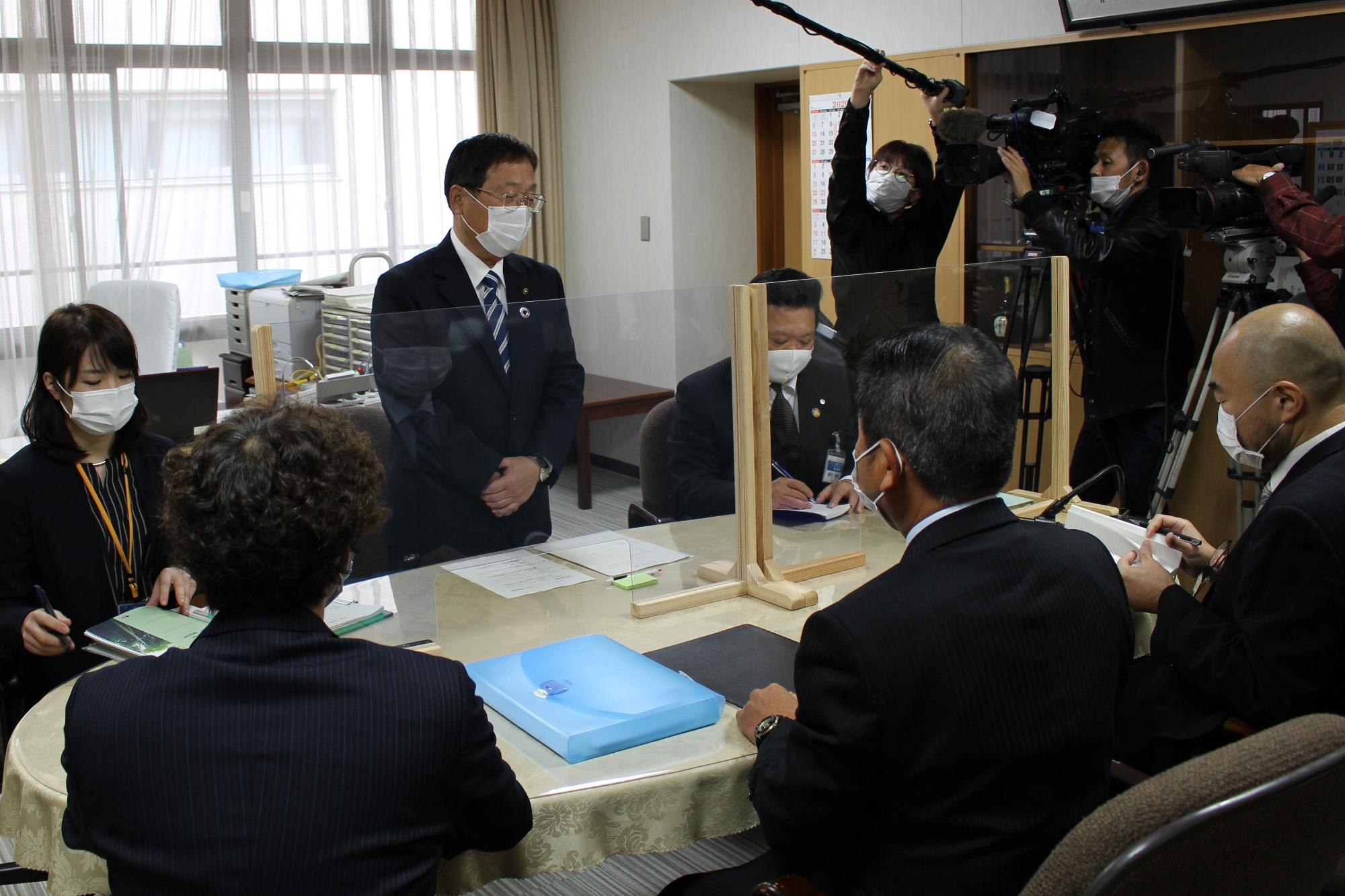 立って話すスーツ姿の男性と楕円形のテーブルに座っているスーツ姿の男女たちの写真
