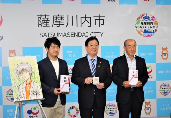 迫田会長、山下代表取締役と並んで写真に写る田中市長の写真