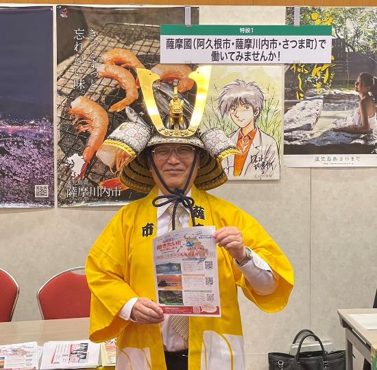 ブースの前で兜を被りPRチラシを手に写真に写る田中市長
