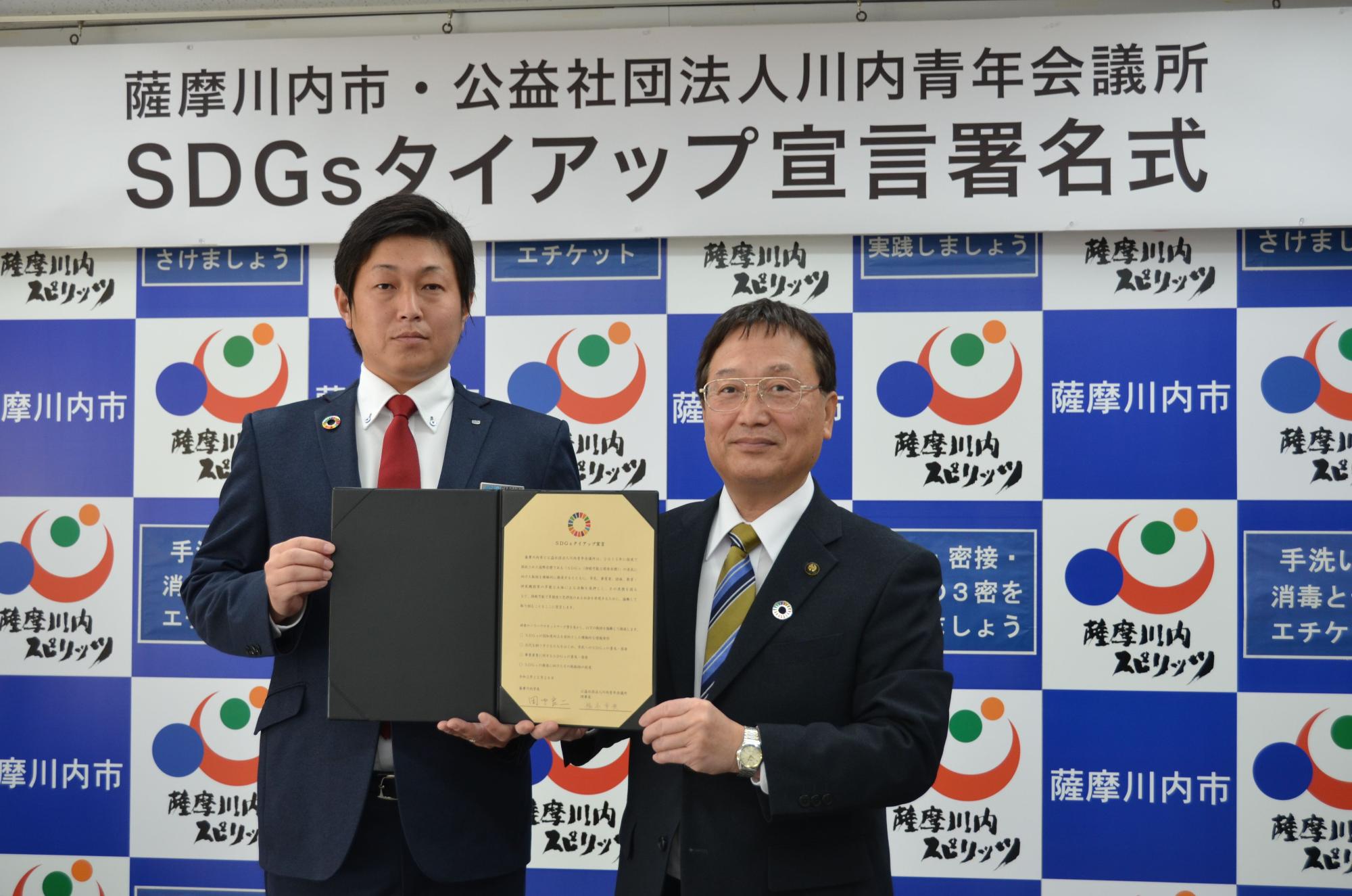 署名した書類を掲げて見せるスーツ姿の男性二人の写真