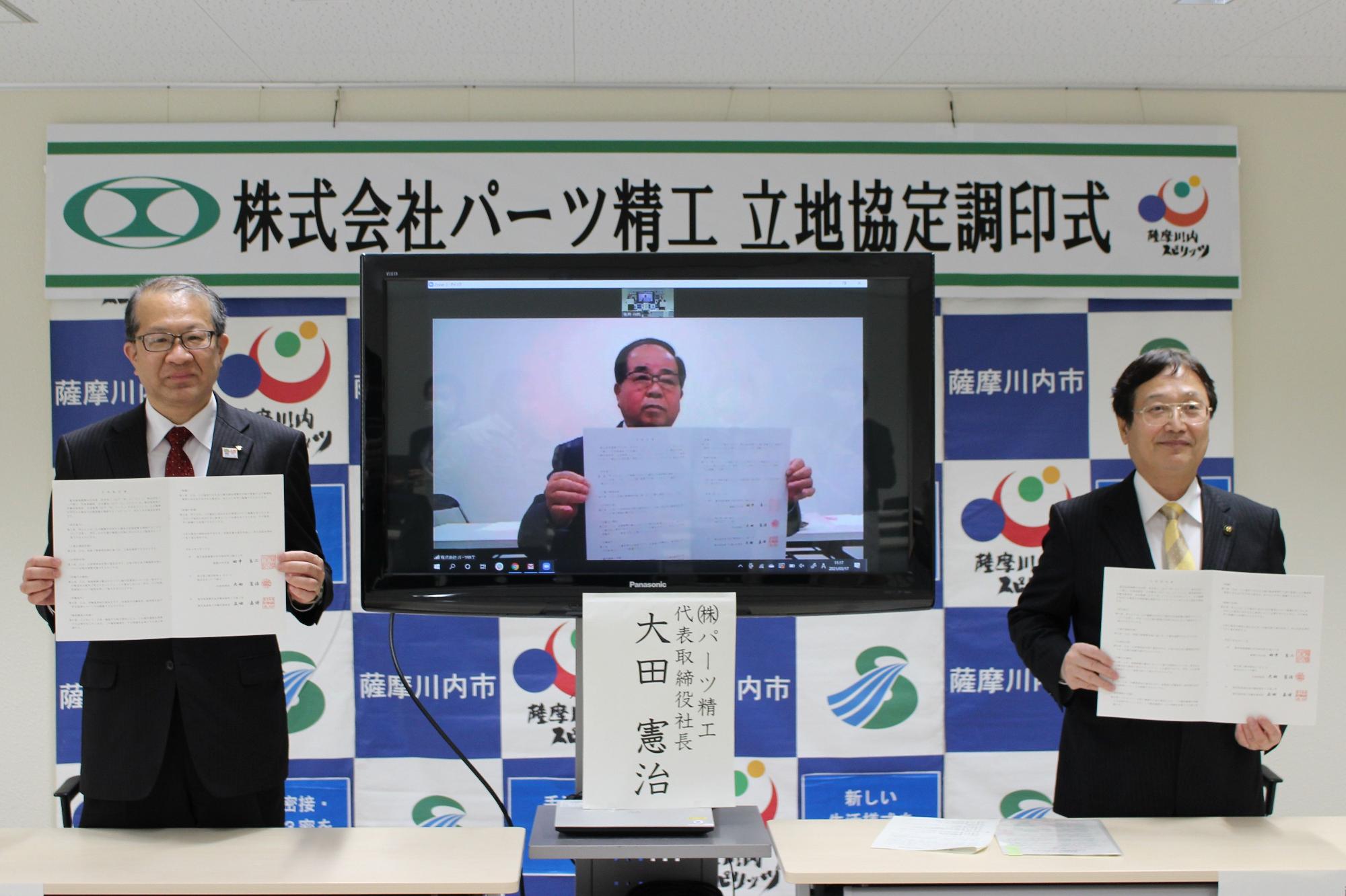 立地協定調印式の証書を見せているテレビ参加の社長と市長の写真