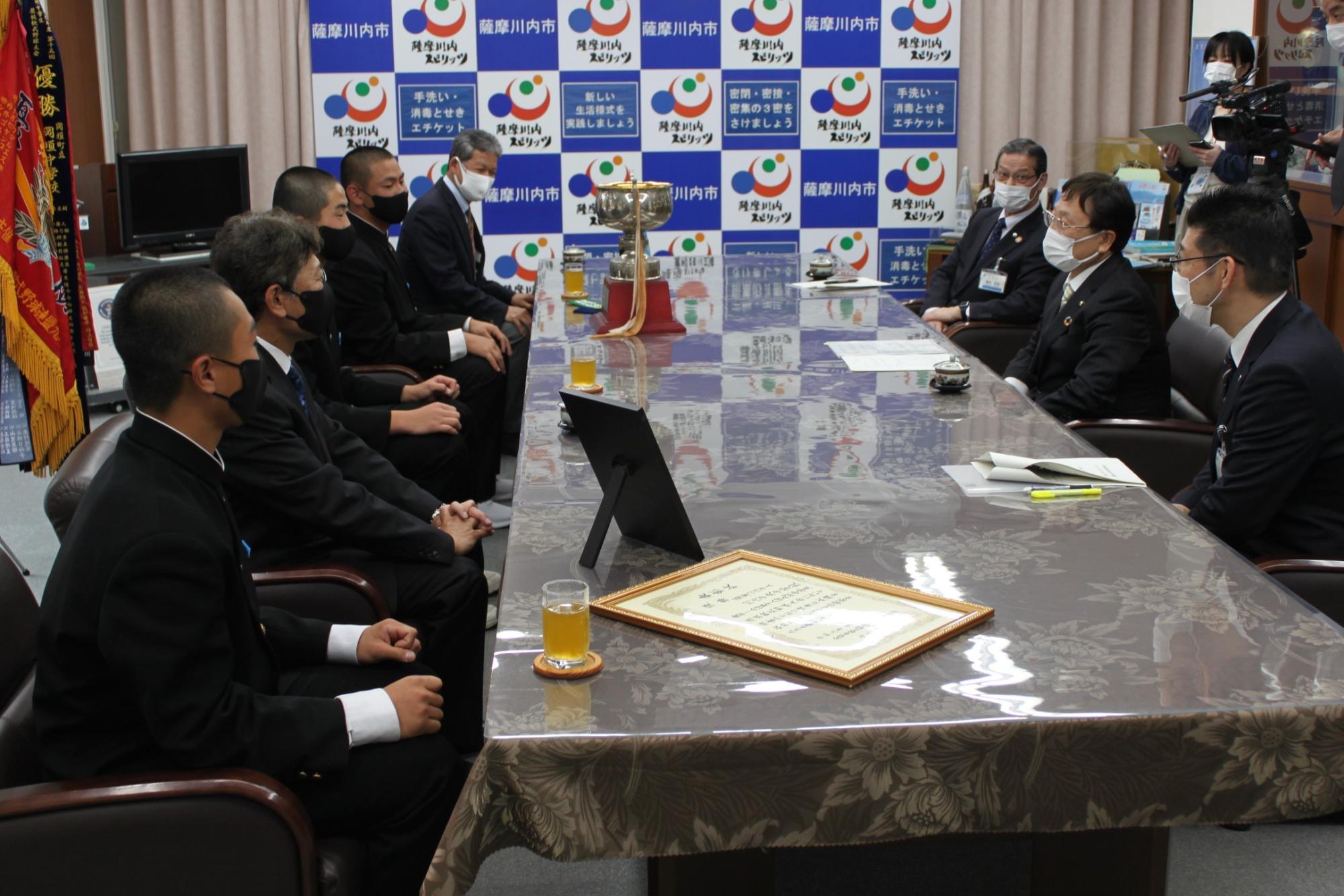 トロフィーが置かれたテーブルを挟んで話をしている、市長ら3人と、監督と4人の選手代表の写真