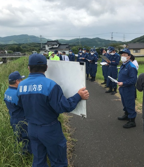 大きな資料を掲げて見せている青い服の男性とそれを聞いているほかの男性たちの写真