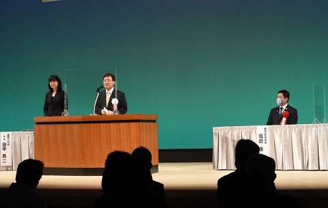記念式典会場の壇上でスピーチするスーツ姿の男性の写真