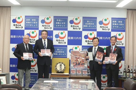 川内大綱引を継承する会の関係者、市長ら4人が映画「大綱引の恋」のポスターを中心に、並んでDVDを手にしている写真