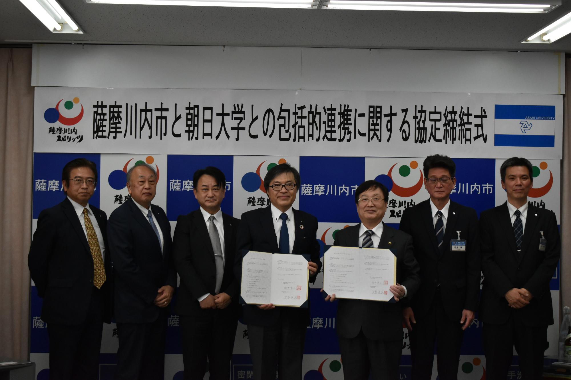 薩摩川内市と朝日大学との包括的連携に関する協定締結式と書かれた垂れ幕の前で調印書を開いている市長と関係者6人の記念写真