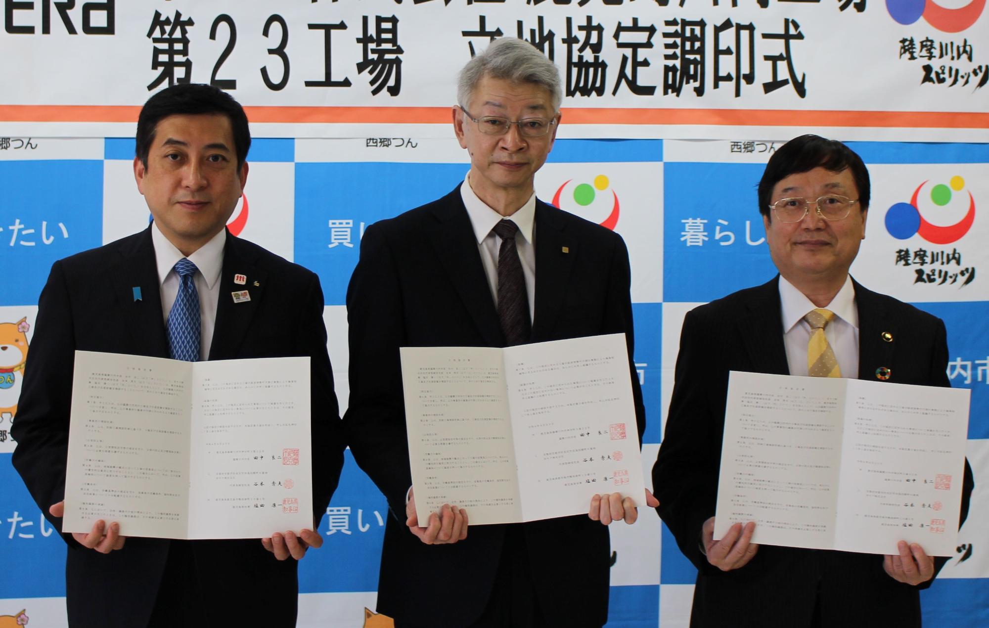 第23工場立地協定調印式と書かれた垂れ幕の前で調印書を開いている市長と鹿児島県知事と京セラの関係者の写真