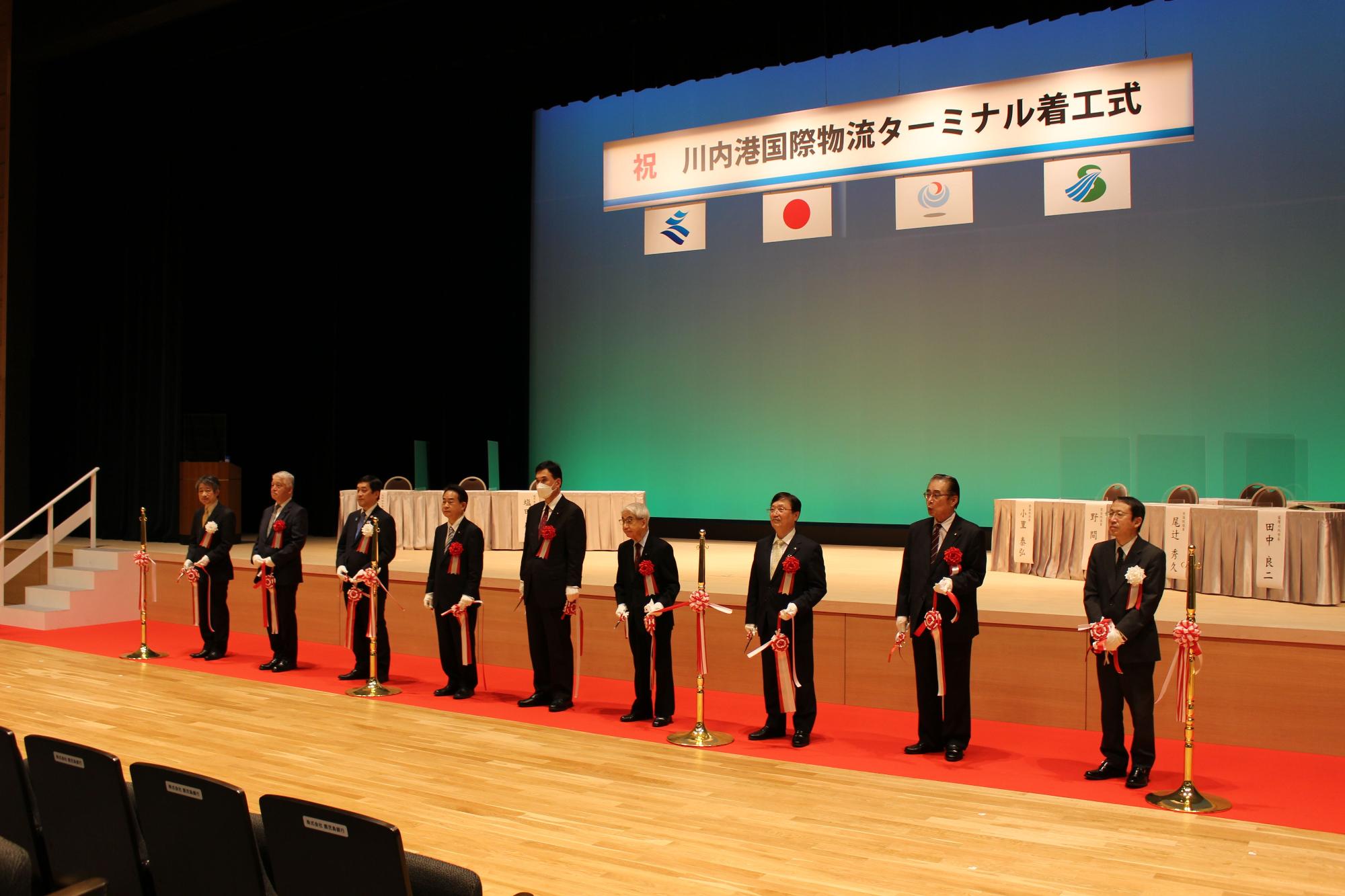 祝、川内港国際物流ターミナル着工式と書かれた幕のもと、テープカットを行った市長ら関係者9人の写真