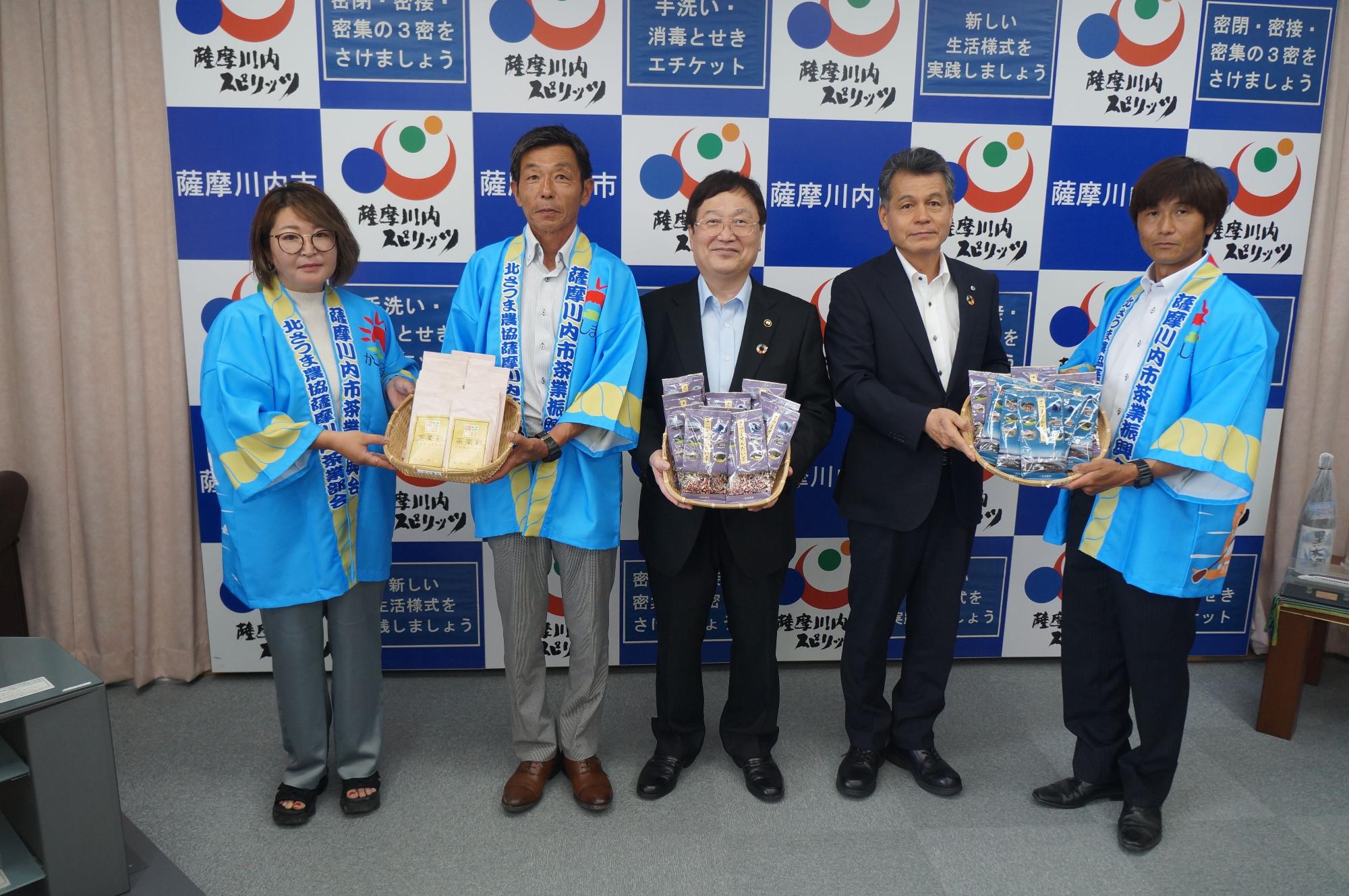 緑茶のパッケージが入ったかごを持つ田中市長とスーツ姿の男性たち、水色の法被を着た男女の写真