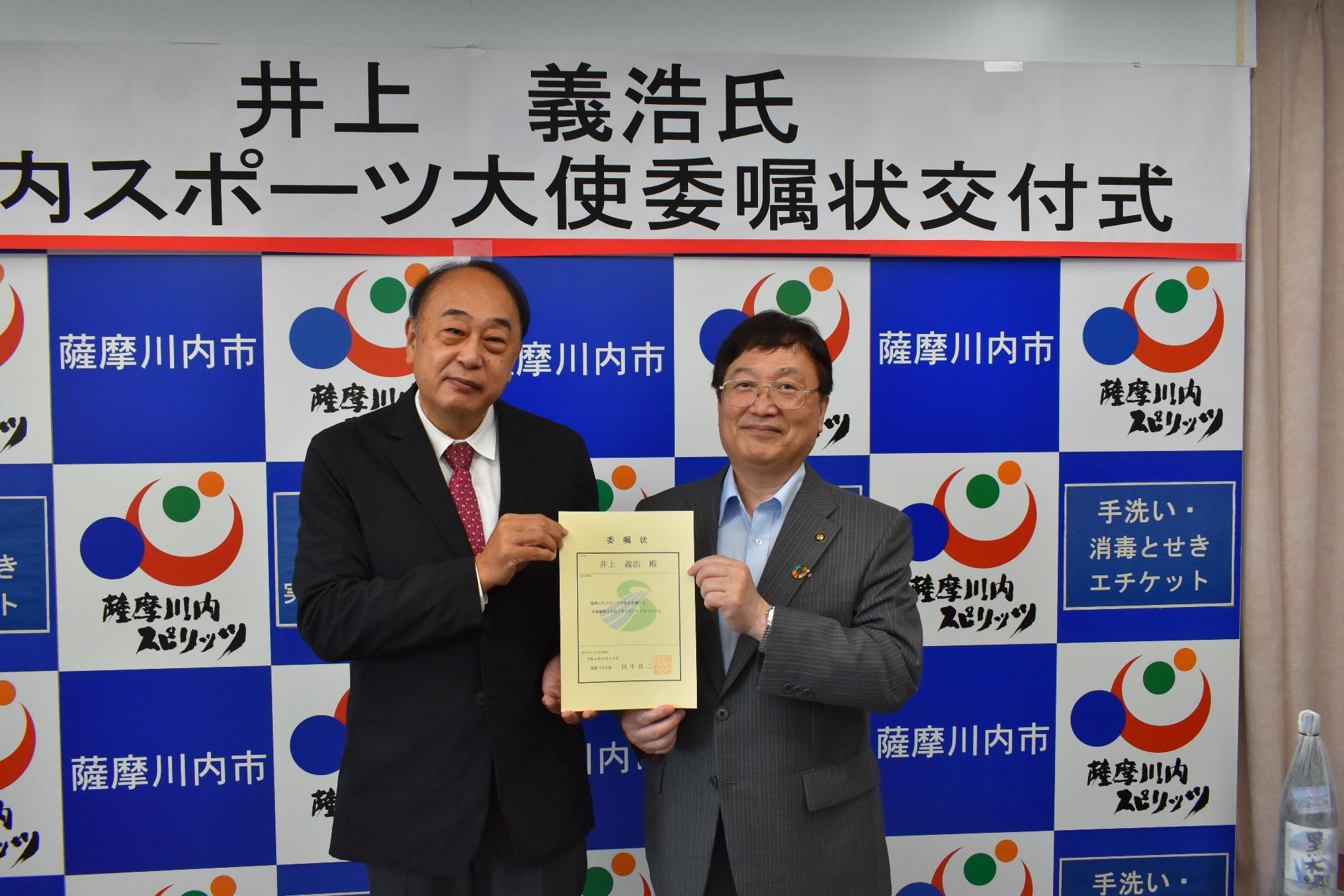 委嘱状を一緒に持ちながら笑う田中市長と井上さんの写真