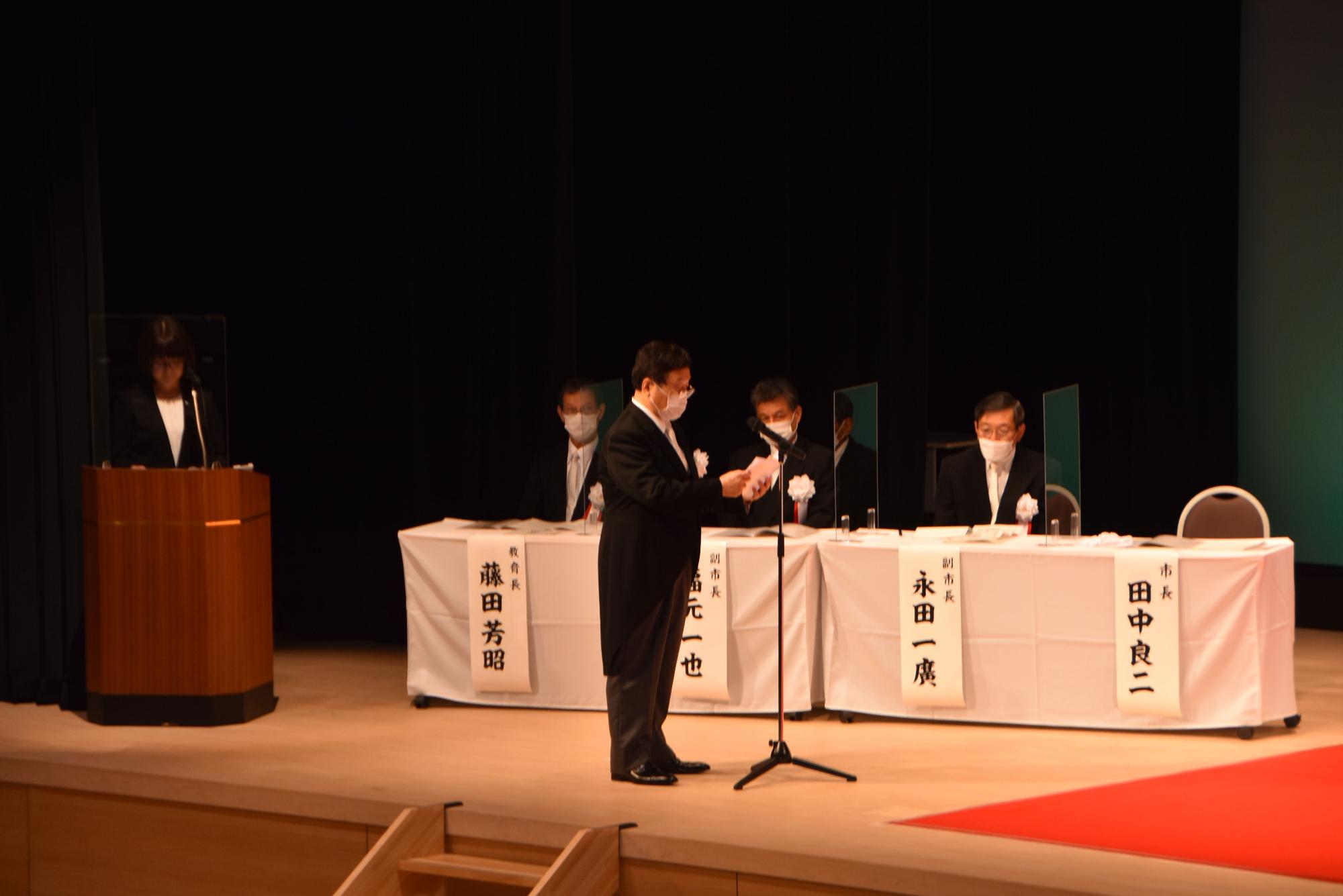壇上で話すスーツ姿の男性とその横に座る副市長たちの写真