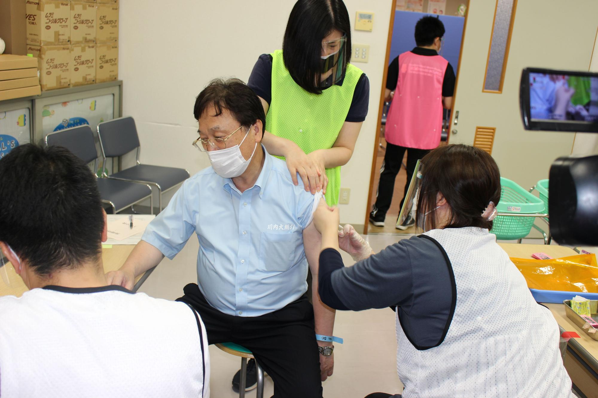 黄緑のビブスを着た女性に肩を押さえられ看護師に注射をされる青いシャツの男性の写真