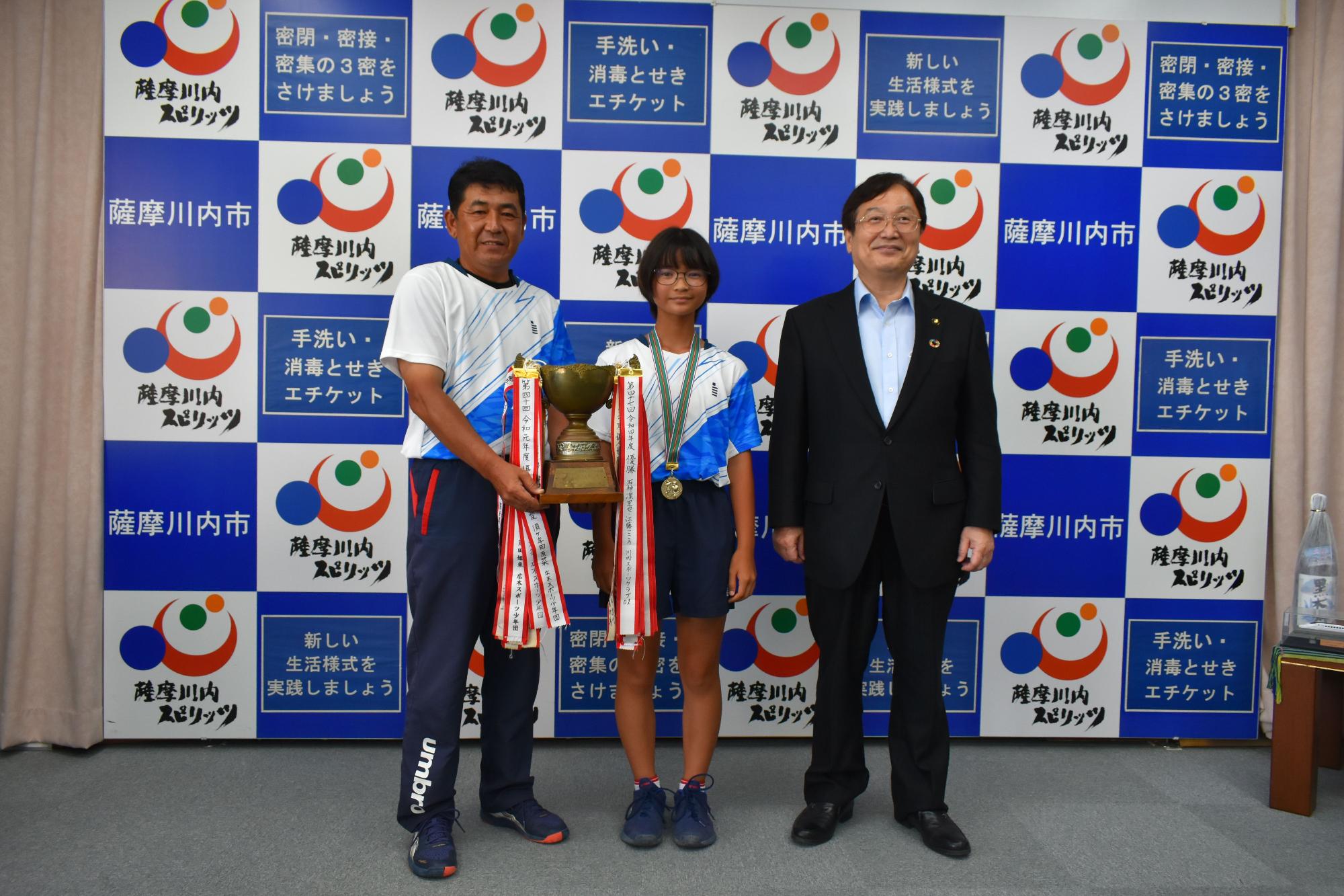 小学生とその両脇に立つスーツ姿とTシャツ姿の男性たちの写真