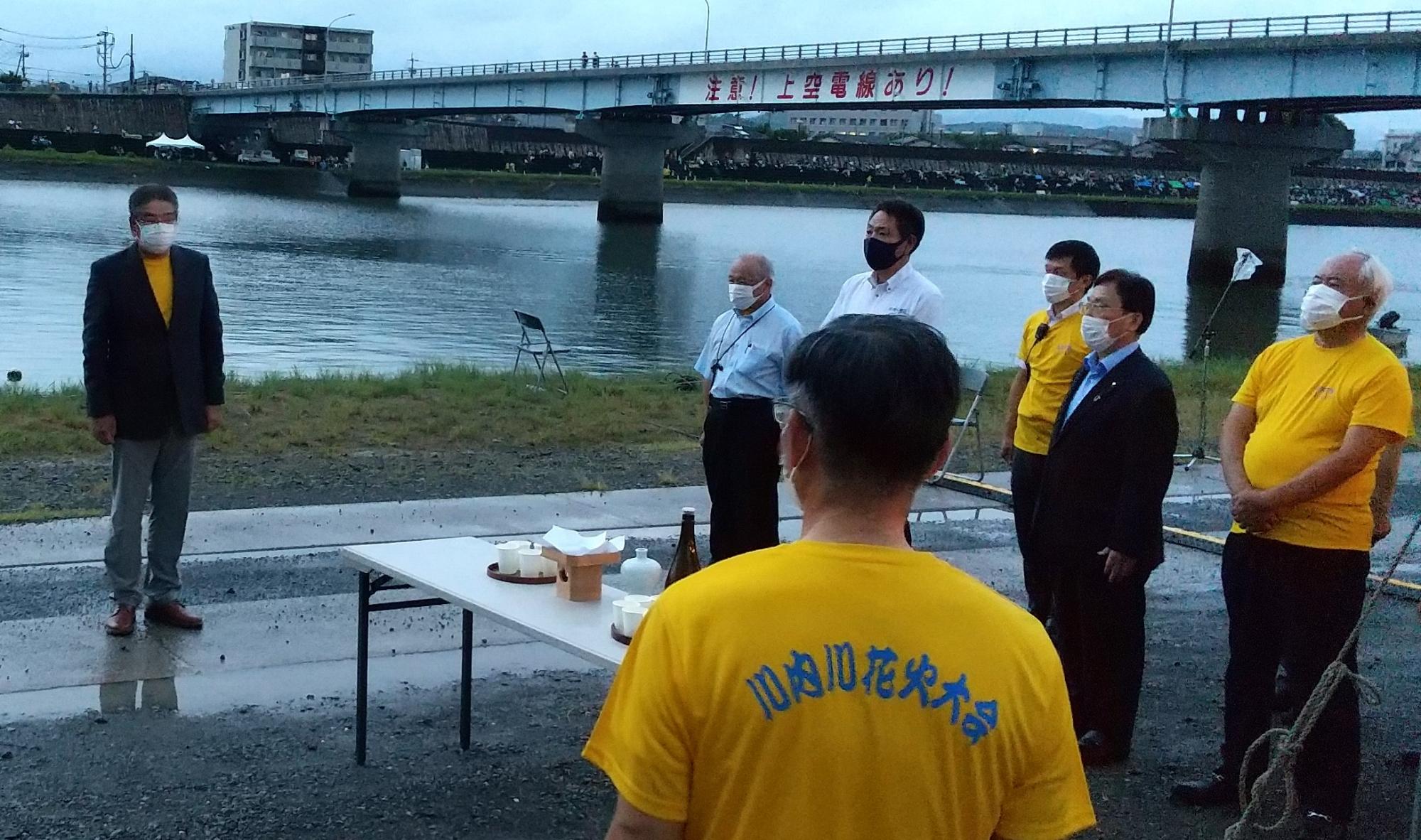 雨上がりの川の前で話すスーツ姿の男性と川内川花火大会のTシャツを着た男性たちの写真