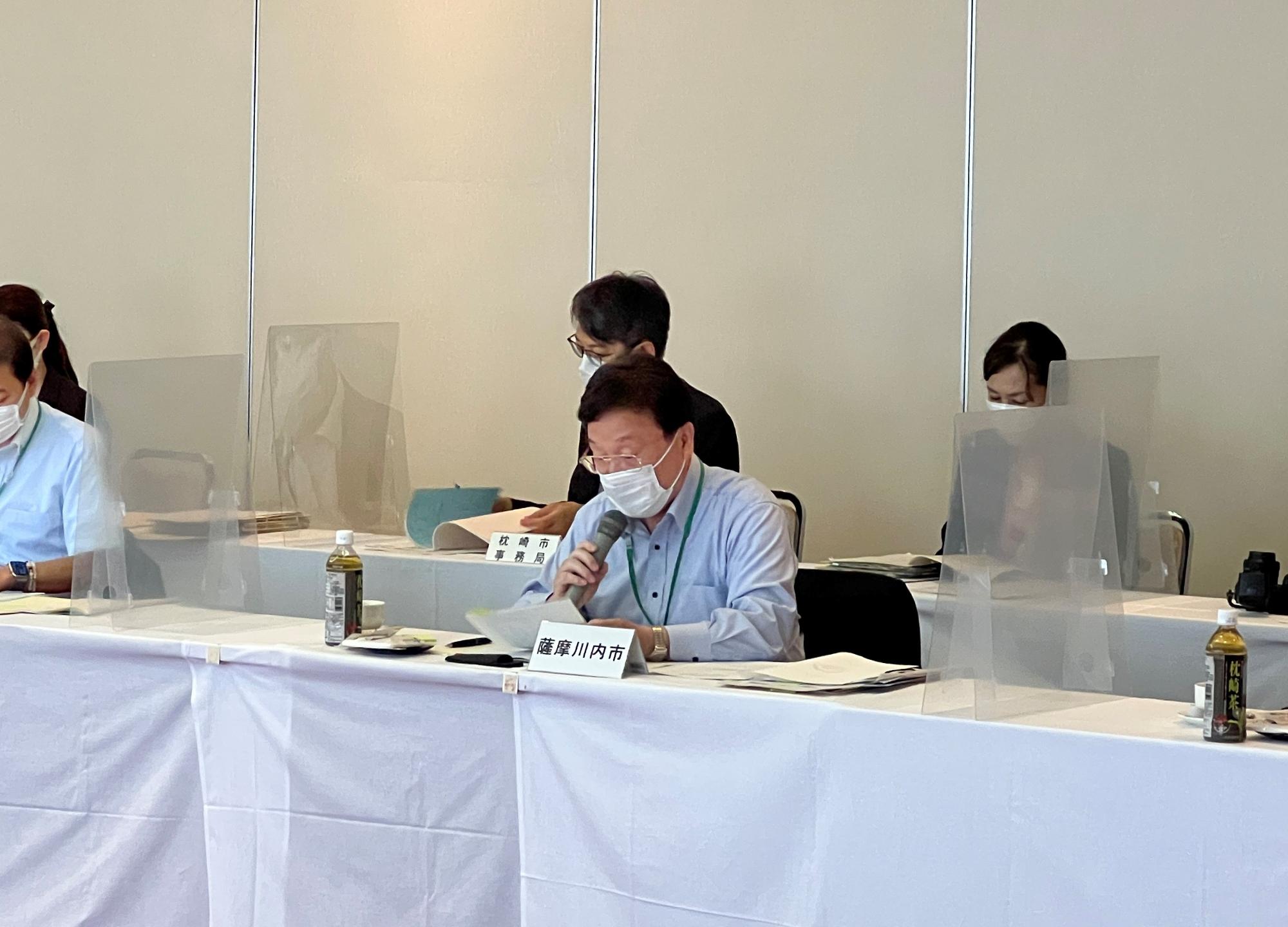 薩摩川内市の席札が置かれたテーブルでマイクを持って話す青いシャツの男性の写真
