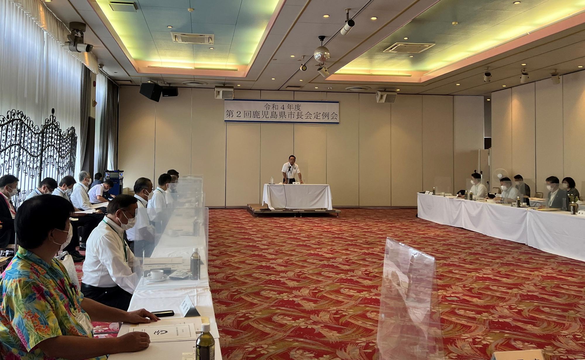 部屋の両側に並んだテーブルに座る大勢の人と中央の席で発言する男性の写真