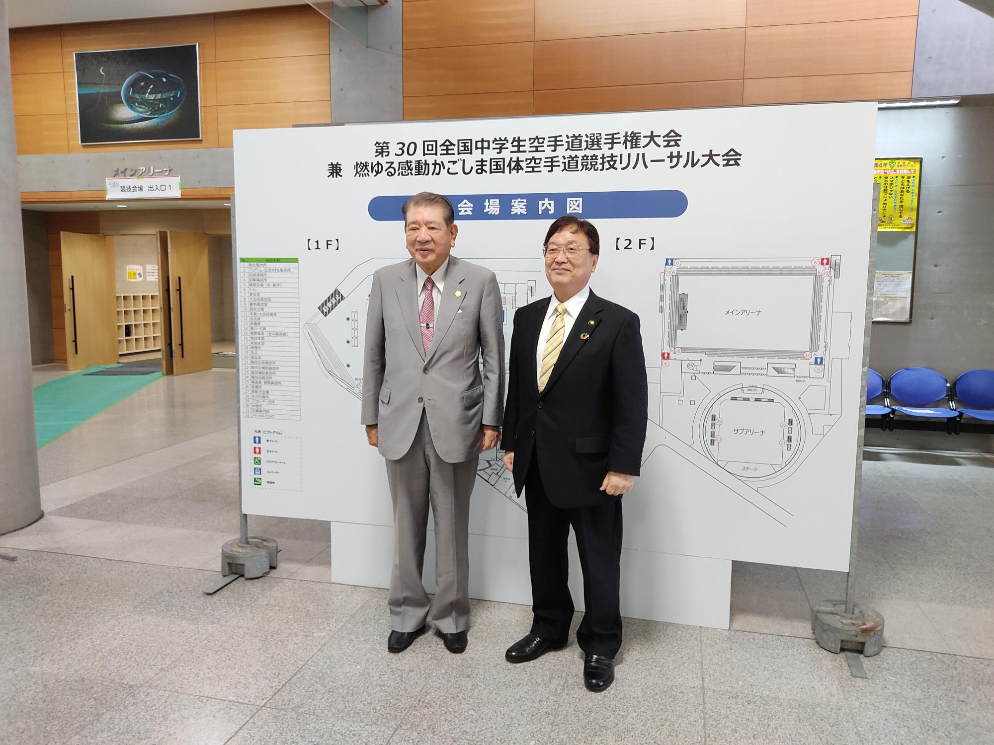 会場案内図の前に並ぶスーツ姿の男性二人の写真