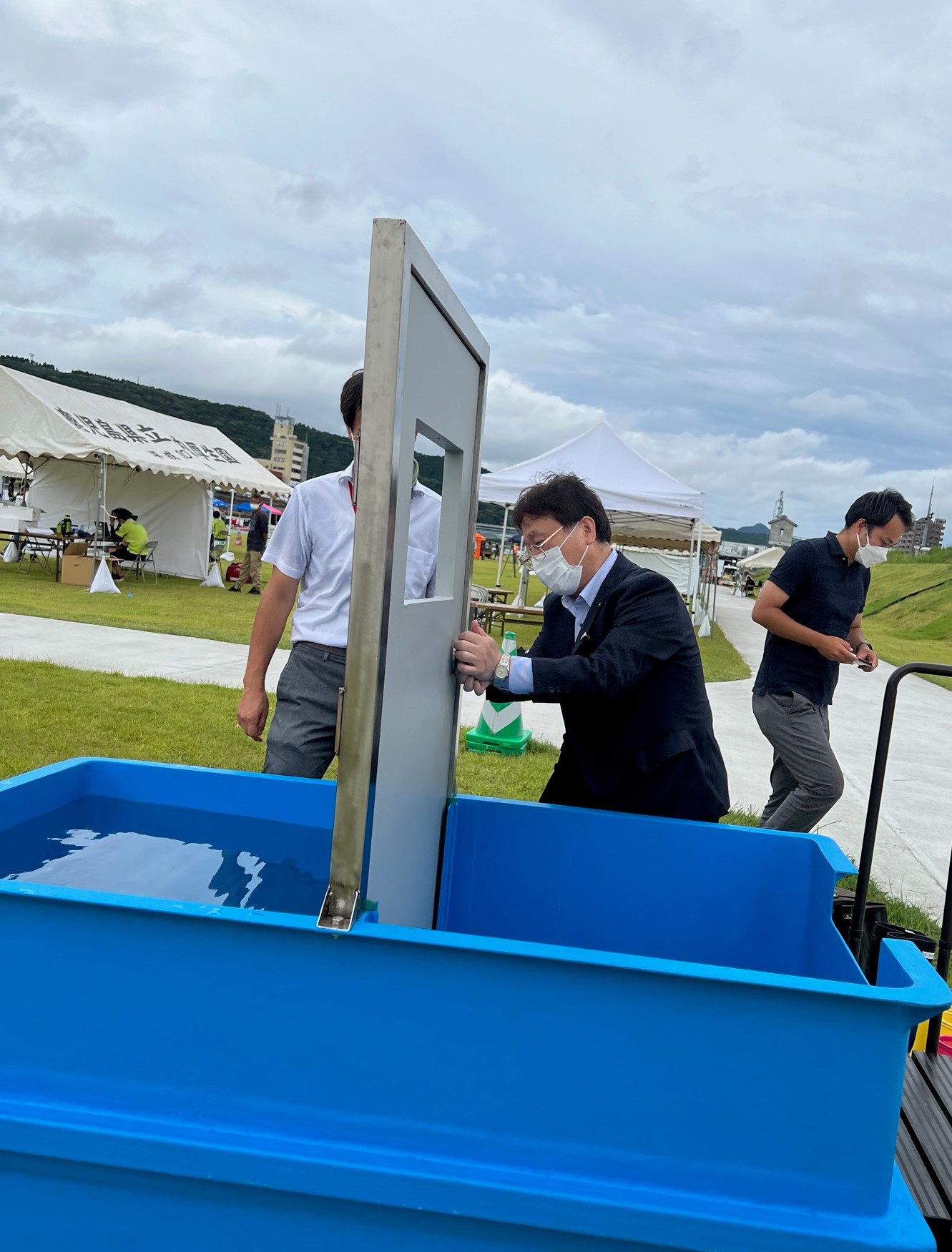 水が張った水槽の中にドアを立ててドアが開くかどうかの実験をする田中市長の写真