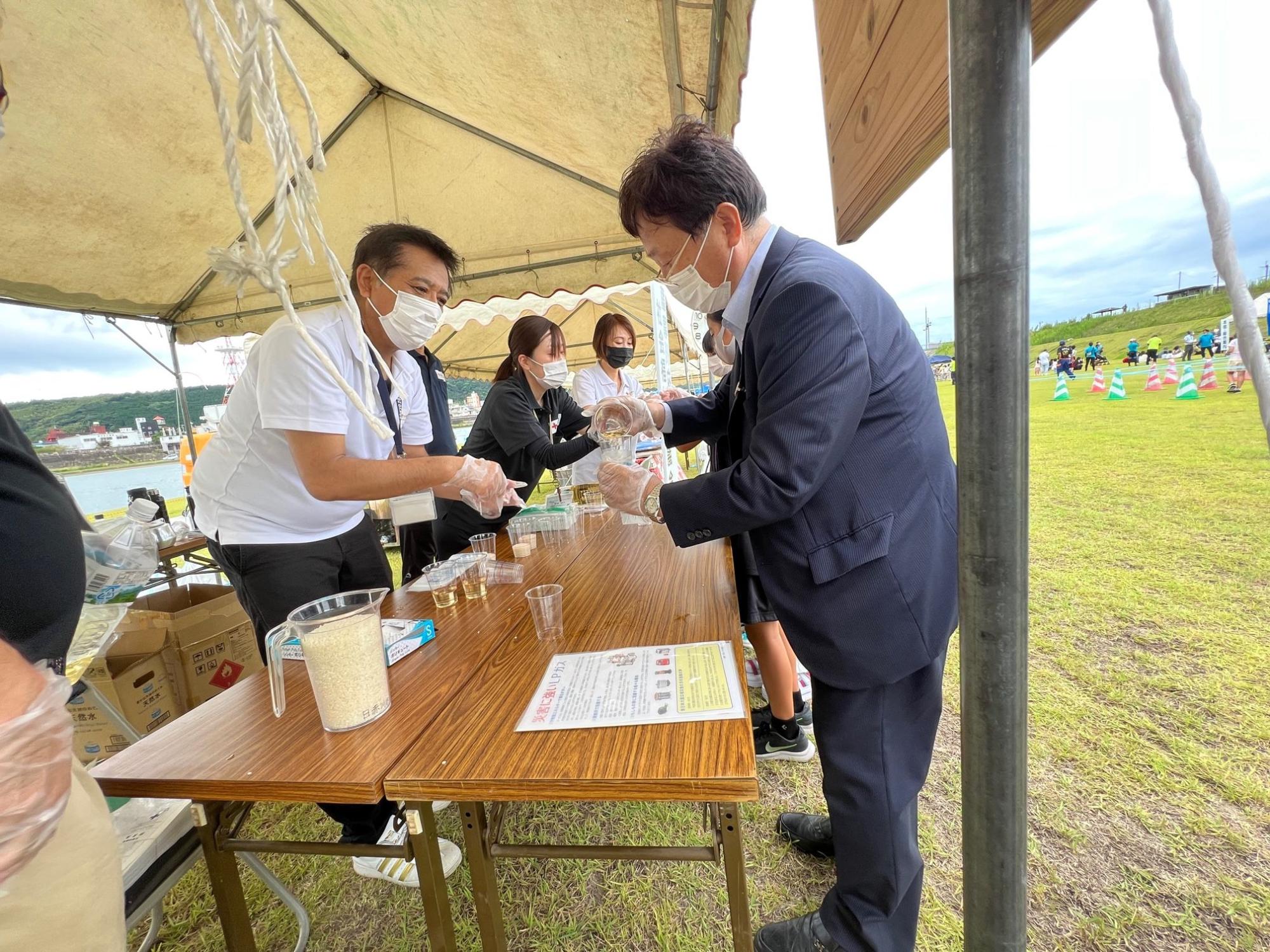手に持ったカップに水をそそぐ田中市長とテーブルの向かい側で市長と話す男性の写真