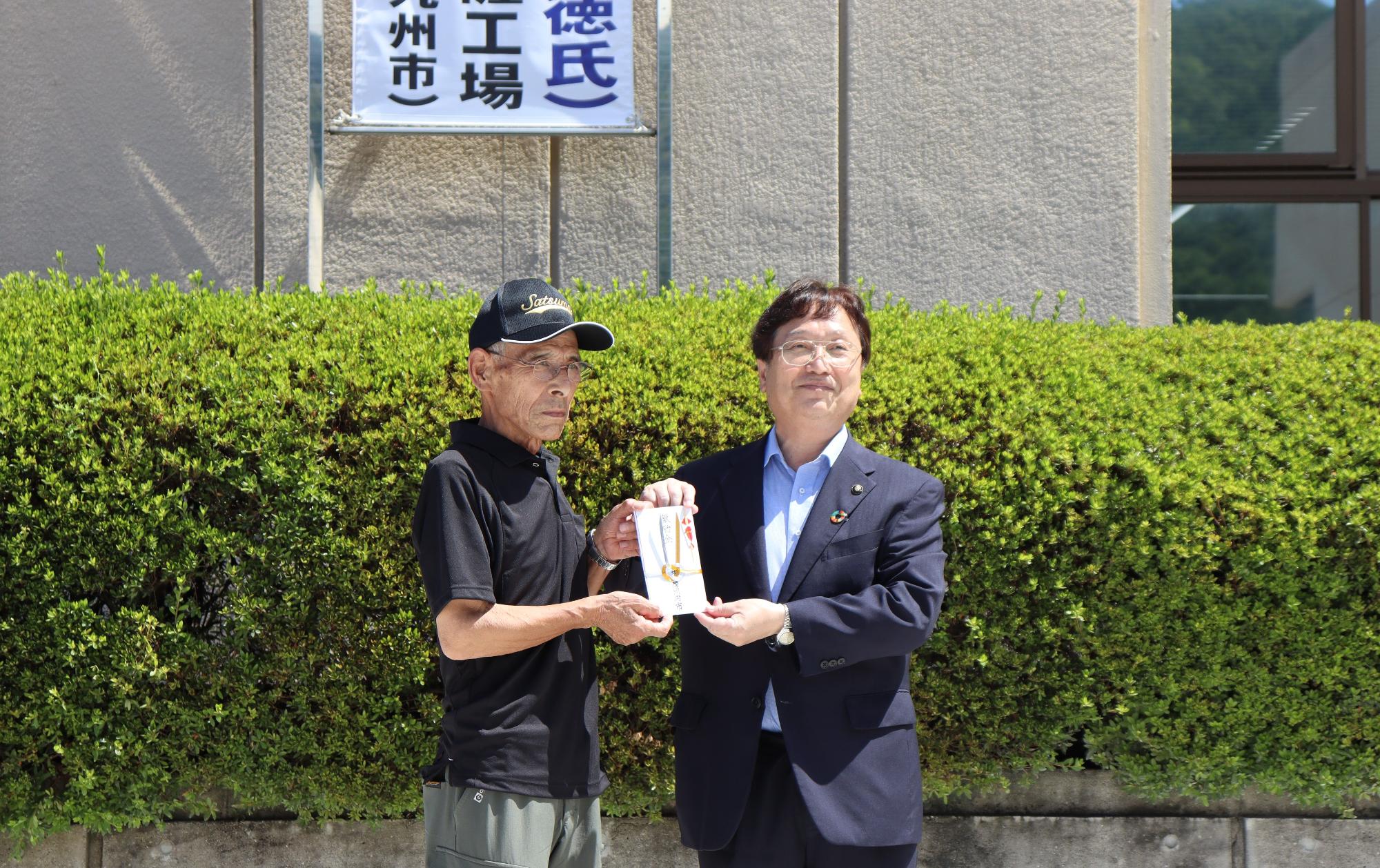 本庁舎前で帽子を被った男性と一緒にのし袋を持つ田中市長の写真