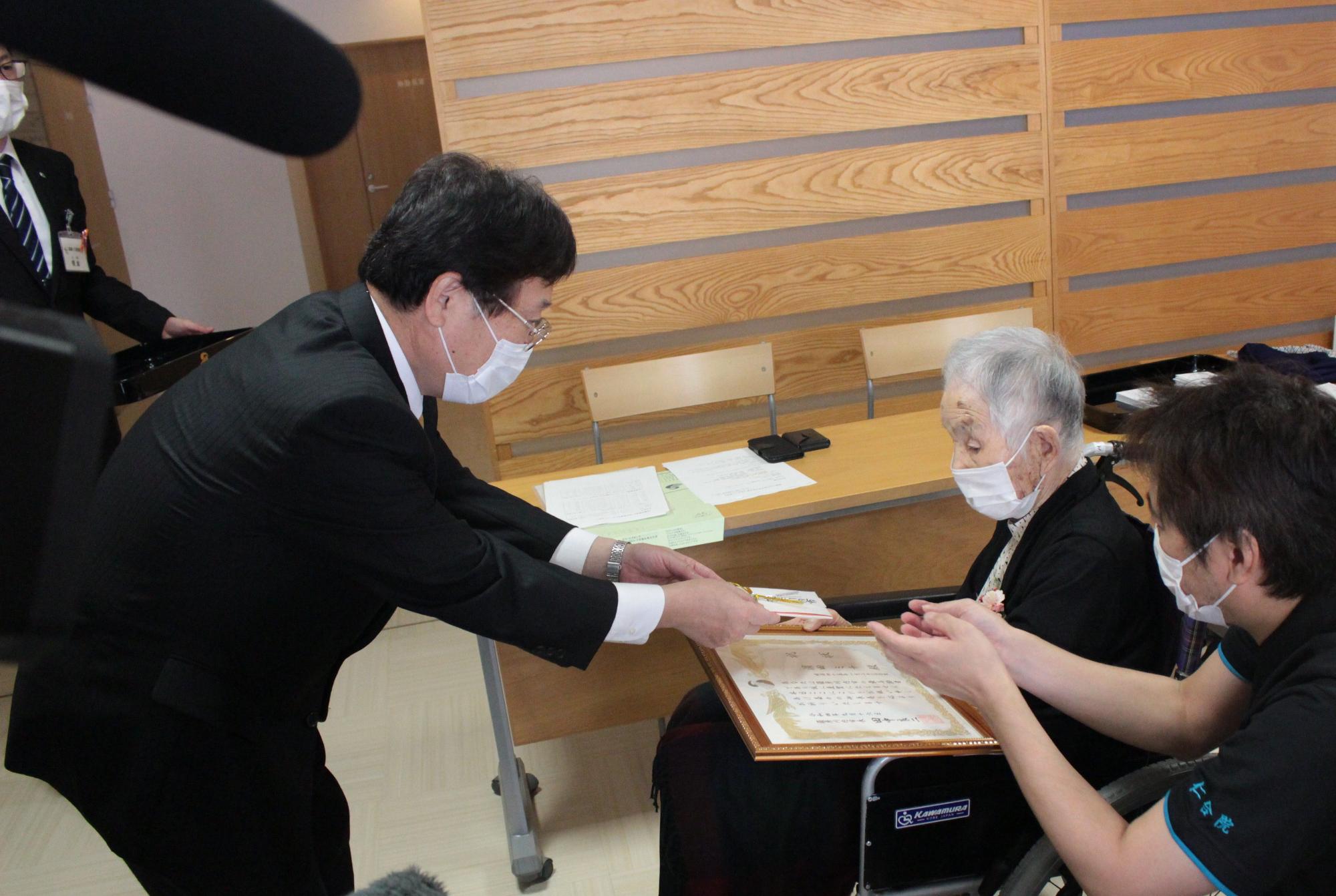 車いすに乗った高齢の女性に賞状を渡す田中市長の写真