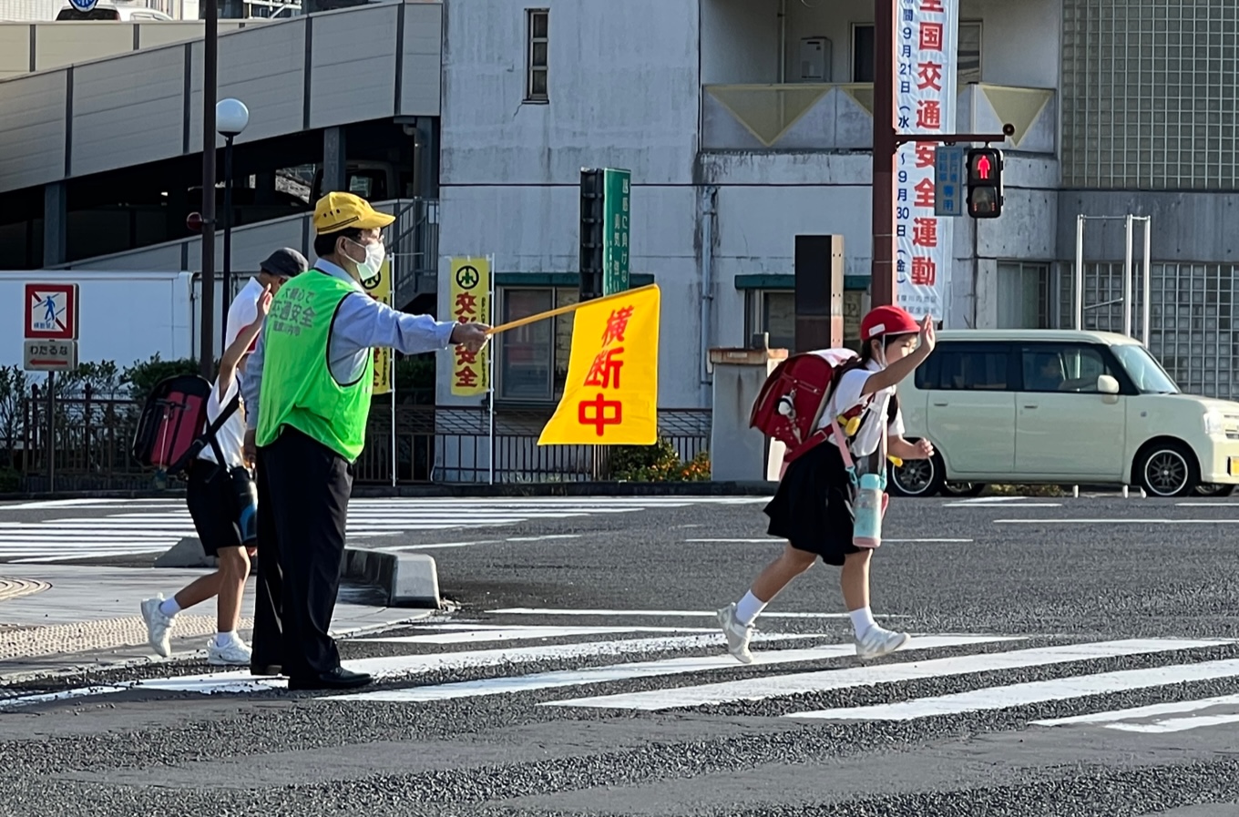 手を上げて横断歩道を渡る小学生たちと横断中と書かれた旗で子供たちを誘導する男性の写真