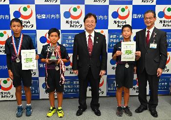 並んで写真に写る隈之城ジュニアソフトテニスクラブの3人と田中市長、藤田教育長の写真