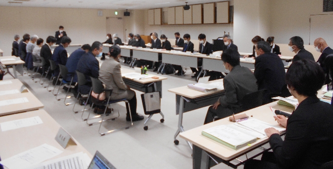 複数の男女が会議室で四角く並べられた長机に向かい合って座り資料を広げている写真