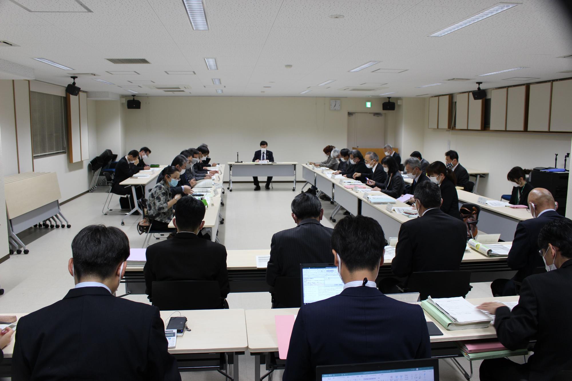 長机が向かい合わせで複数並んだ会議室で30名ほどの男女が資料をひろげ目を通している様子の写真