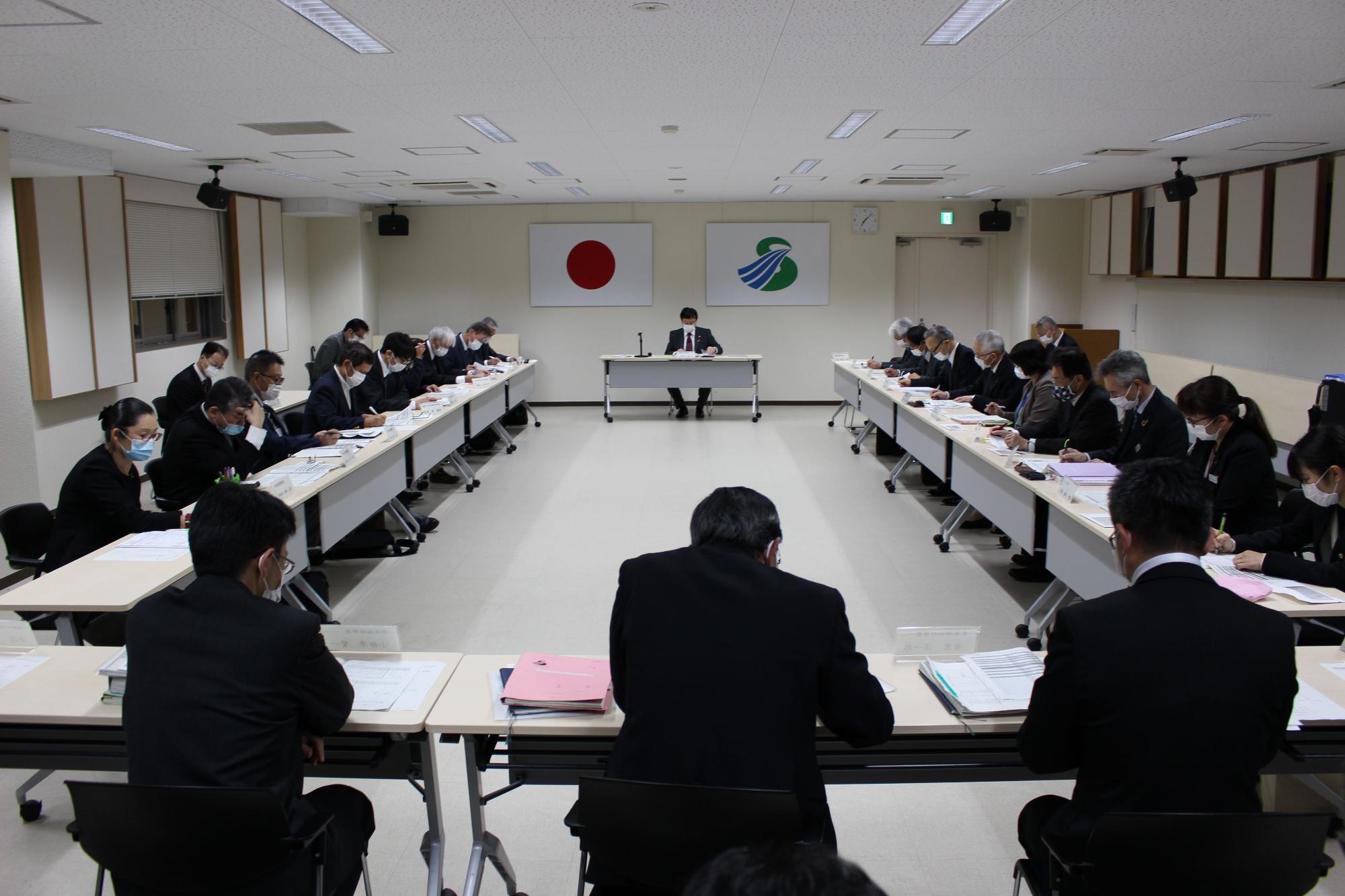 壁に国旗と市旗が飾られた部屋で四角く向かい合って配置された長机にスーツ姿の複数の人が座り資料を見ている写真