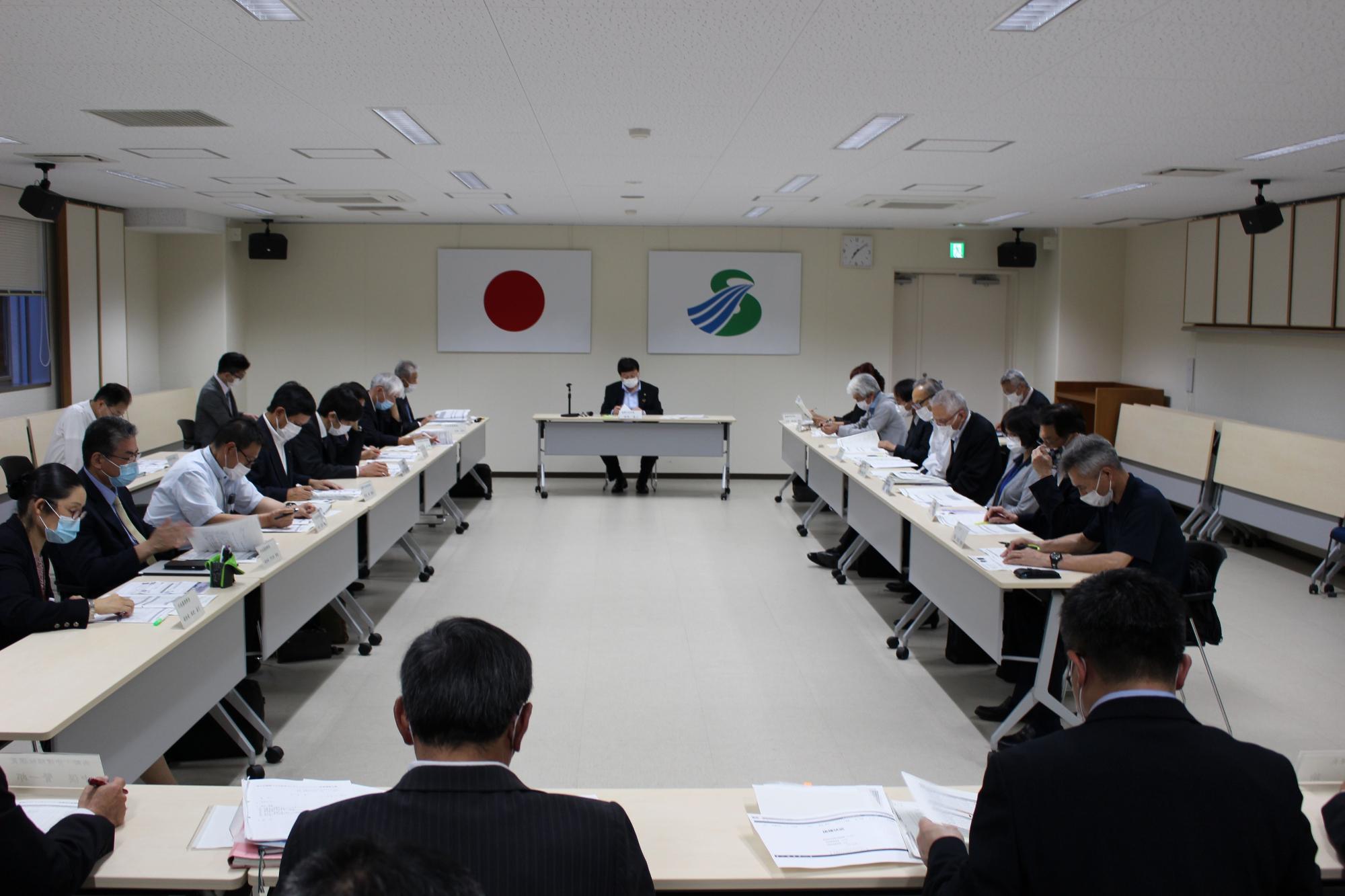 壁に国旗と市旗が飾られた部屋で四角く向かい合って配置された長机に複数の人が座り資料を見ている写真