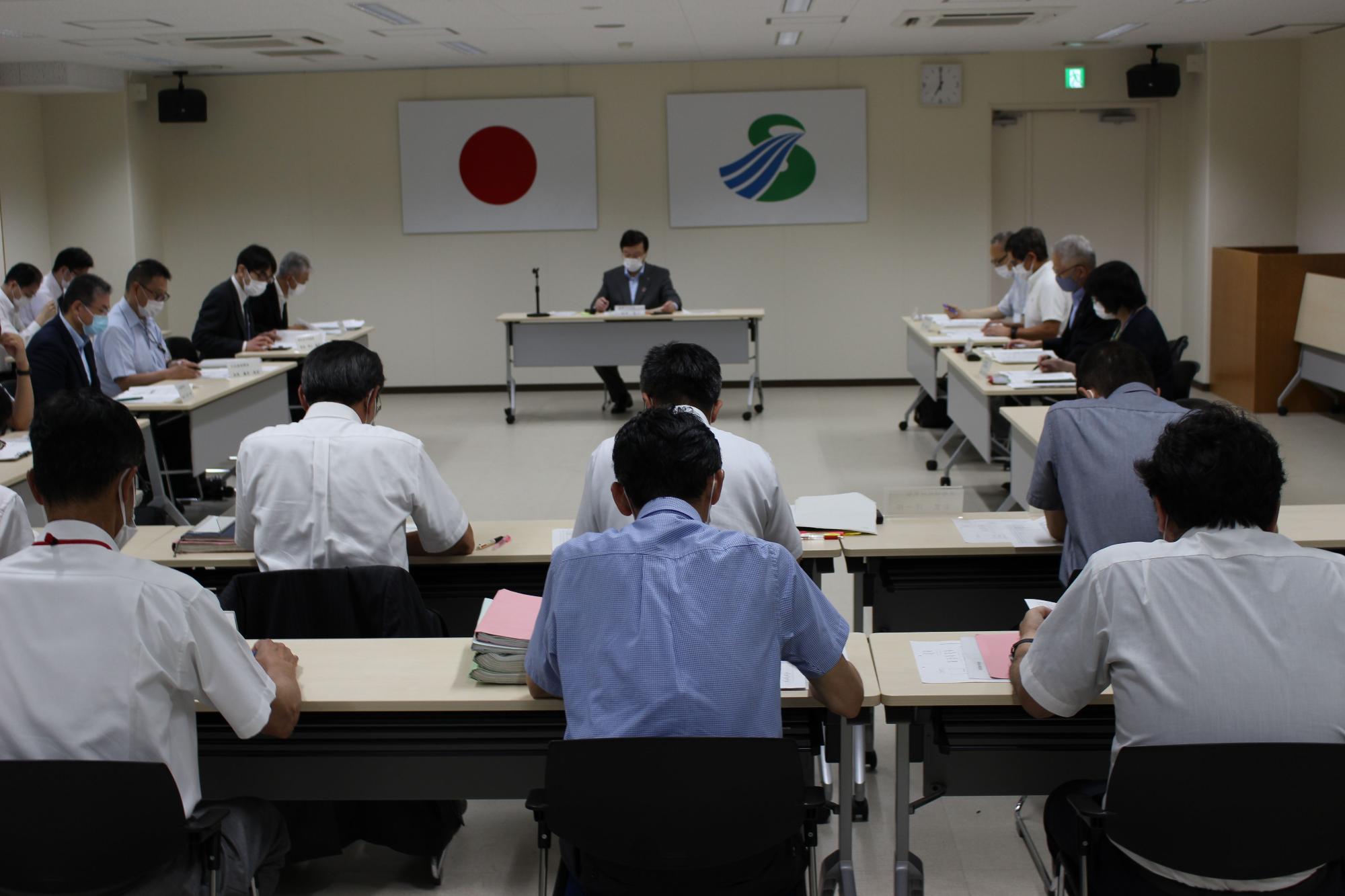 国旗と市旗が飾られた部屋で長机に座り資料を見ながら会議を行っている様子の人々の写真