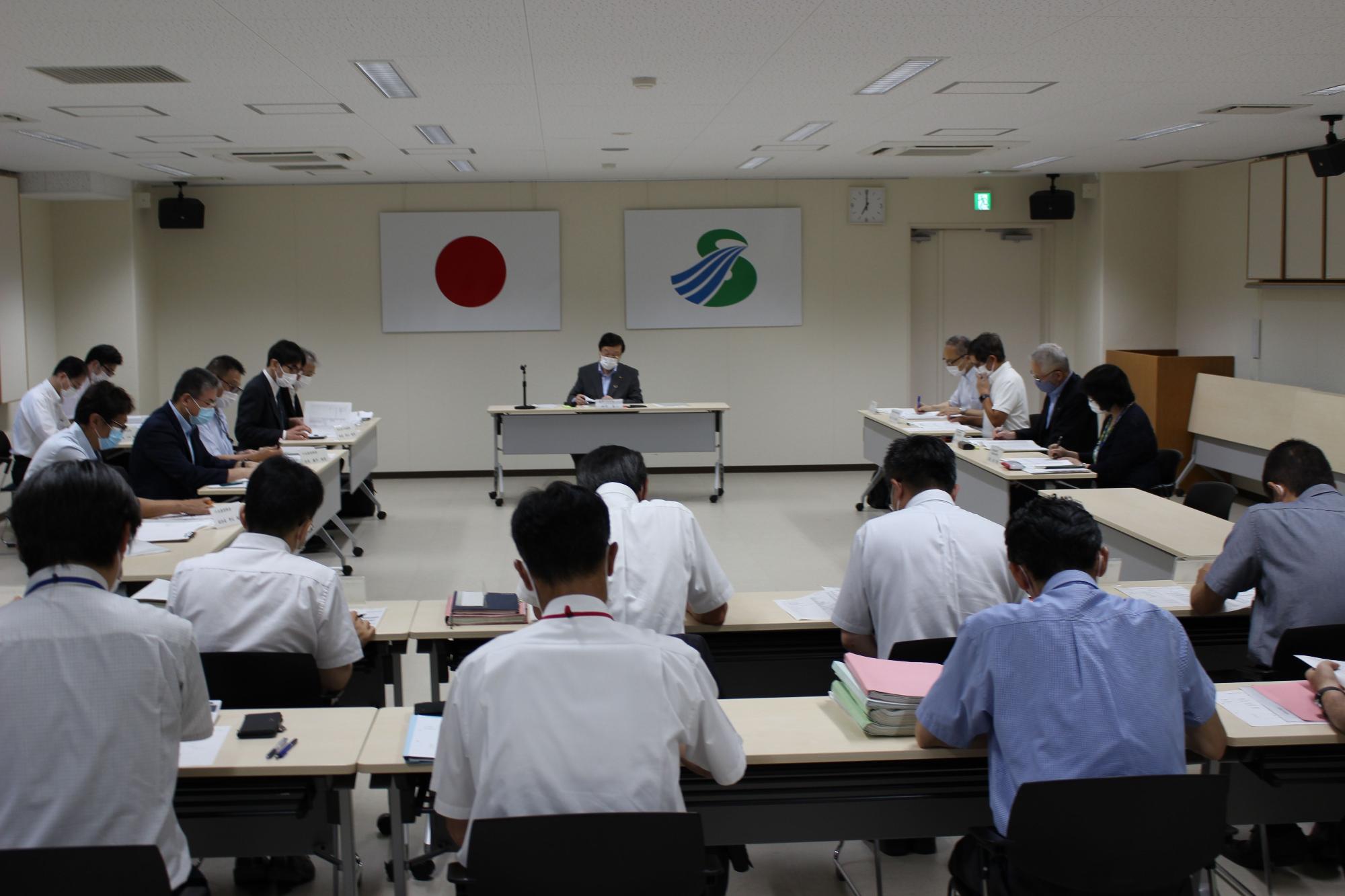 国旗と市旗が飾られた部屋で長机に座り資料を見ながら会議を行っている様子の人々の写真