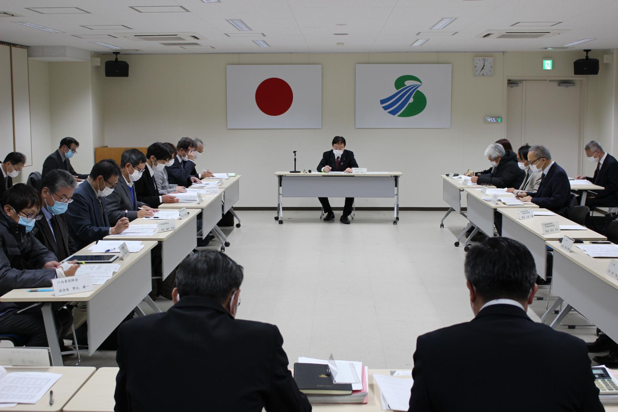 壁に国旗と市旗が飾られた部屋で四角く向かい合って配置された長机に複数の人が座り資料を見ている写真