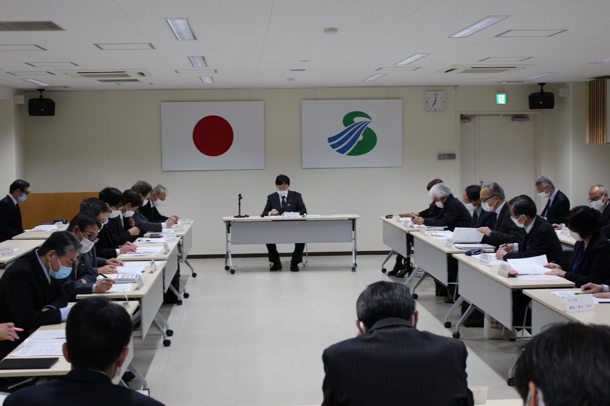 壁に国旗と市旗が飾られた部屋で四角く向かい合って配置された長机にスーツ姿の複数の人が座り資料を見ている写真