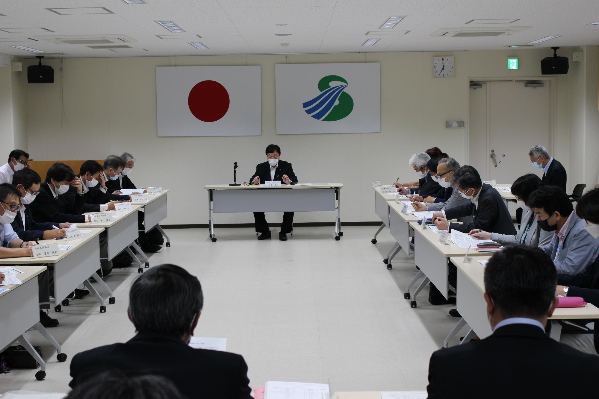 壁に国旗と市旗が飾られた部屋で四角く向かい合って配置された長机にスーツ姿でマスクを付けた複数の人が座り資料を見ている写真