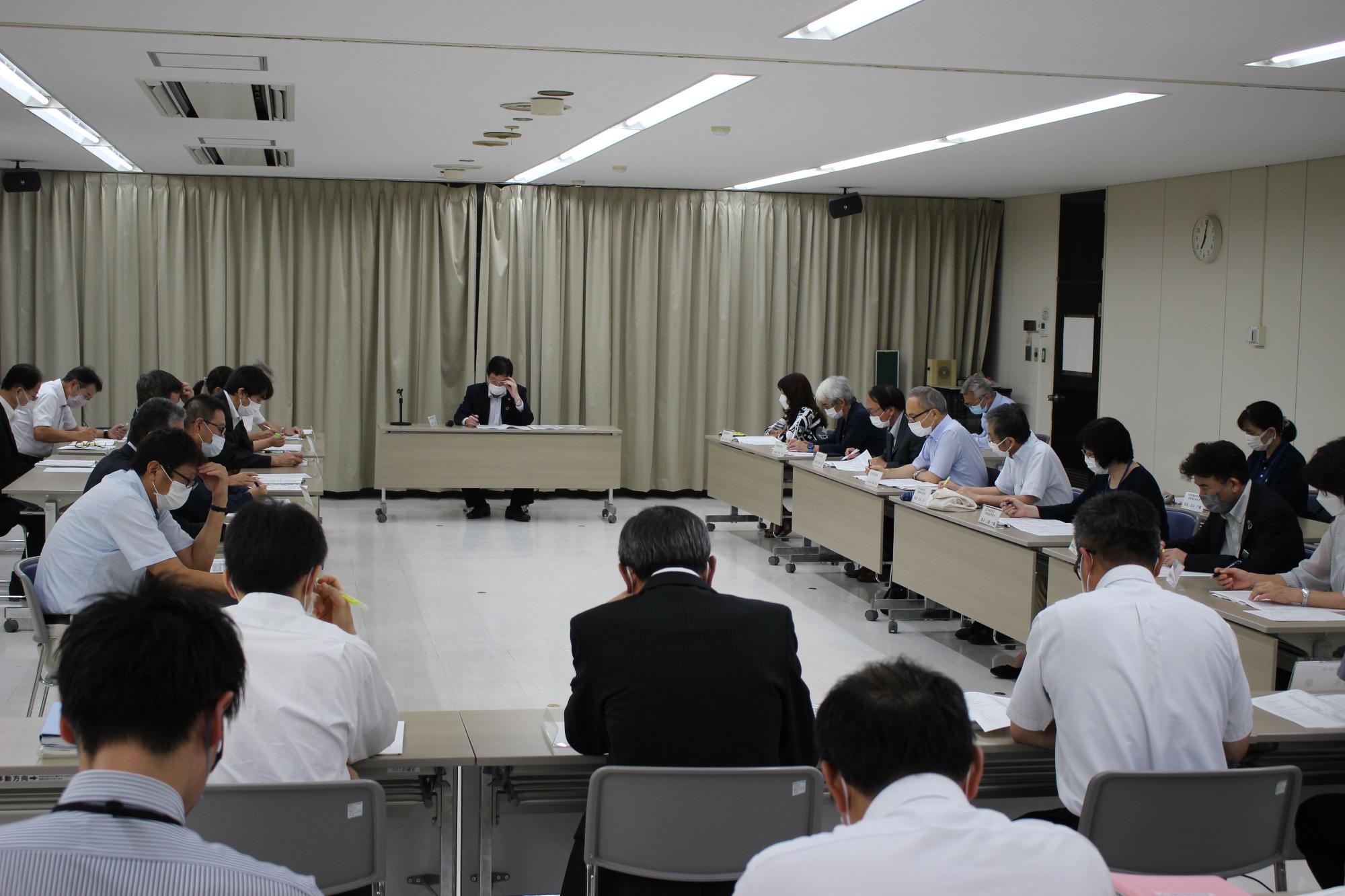 会議室で四角に組んだ長机を囲んで向かい合うように着席した参加者たちが、手元の資料に目を通しながら議事を進めている様子の写真
