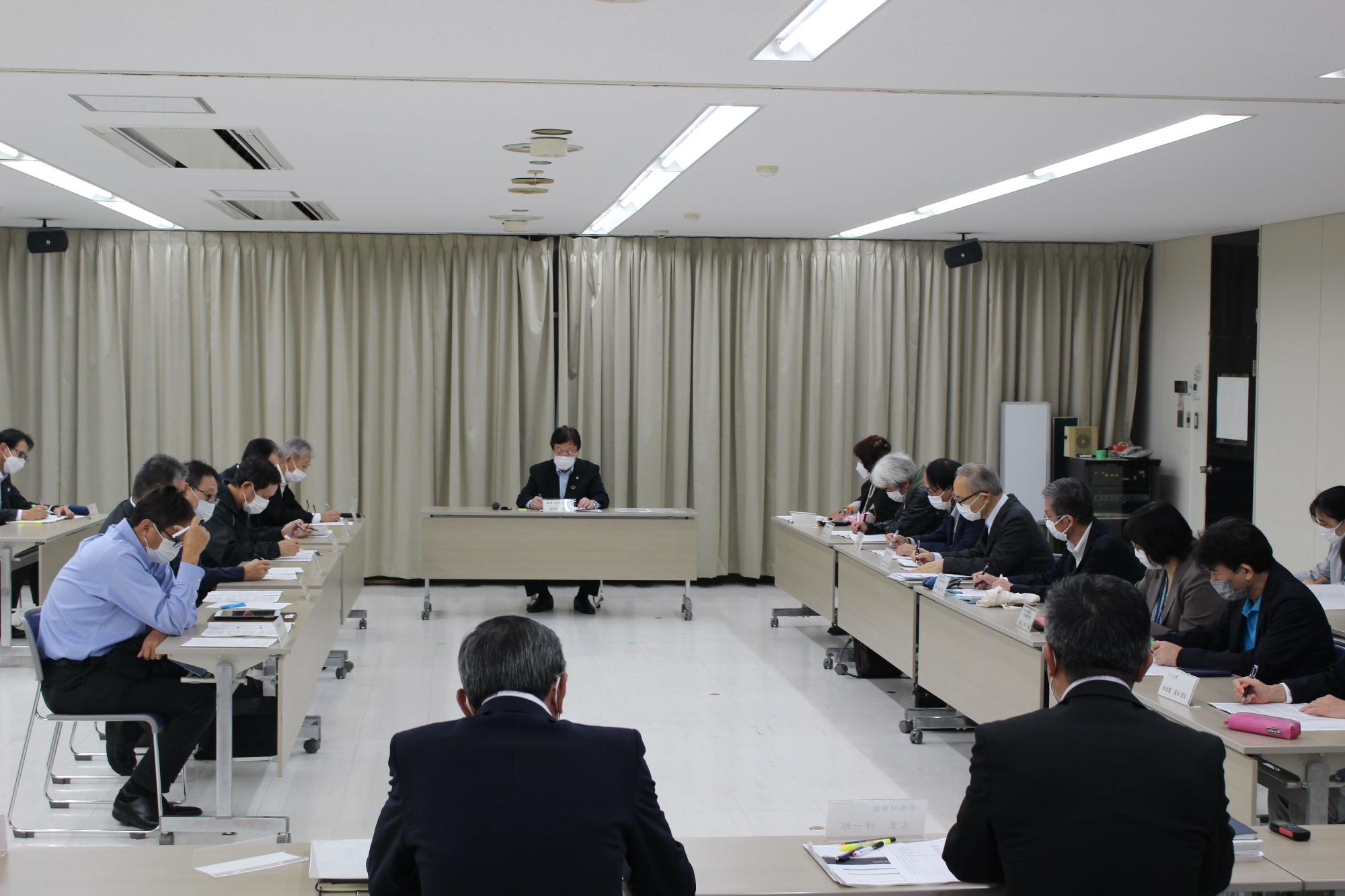 会議室で四角に組んだ長机を囲んで向かい合うように着席した参加者たちが、手元の資料に目を通しながら議事を進めている様子の写真