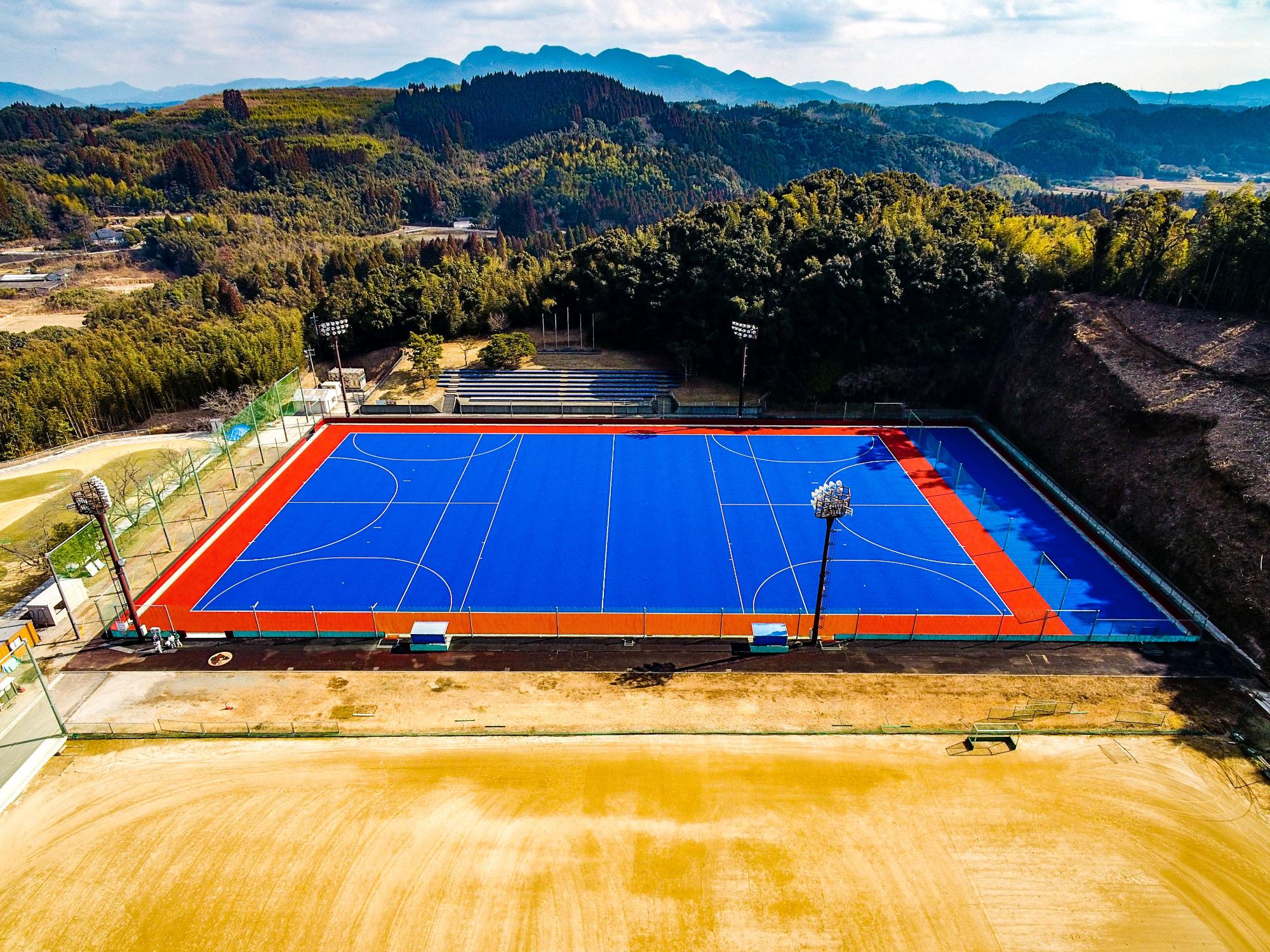 山々に囲まれたグラウンドの隣にある青いコートを上から撮影した写真