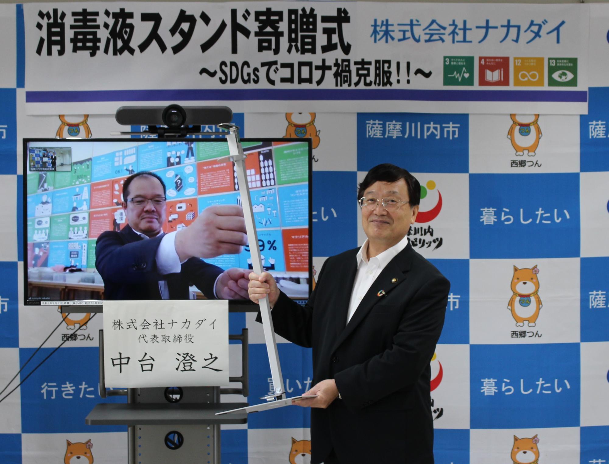 「消毒液スタンド寄贈式~SDGsでコロナ禍克服!!~ 株式会社ナカダイ」と書かれた横断幕の下で、消毒液スタンドを掲げて持つ田中市長とモニターに映る、株式会社ナカダイ代表取締役 中台 澄之氏の写真