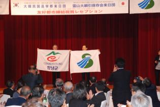 薩摩川内市と昌寧郡のマークの旗を持った男性二人が壇上に立っている写真