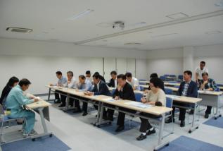 机の並んだ室内でスーツ姿の男性たちが座っている写真