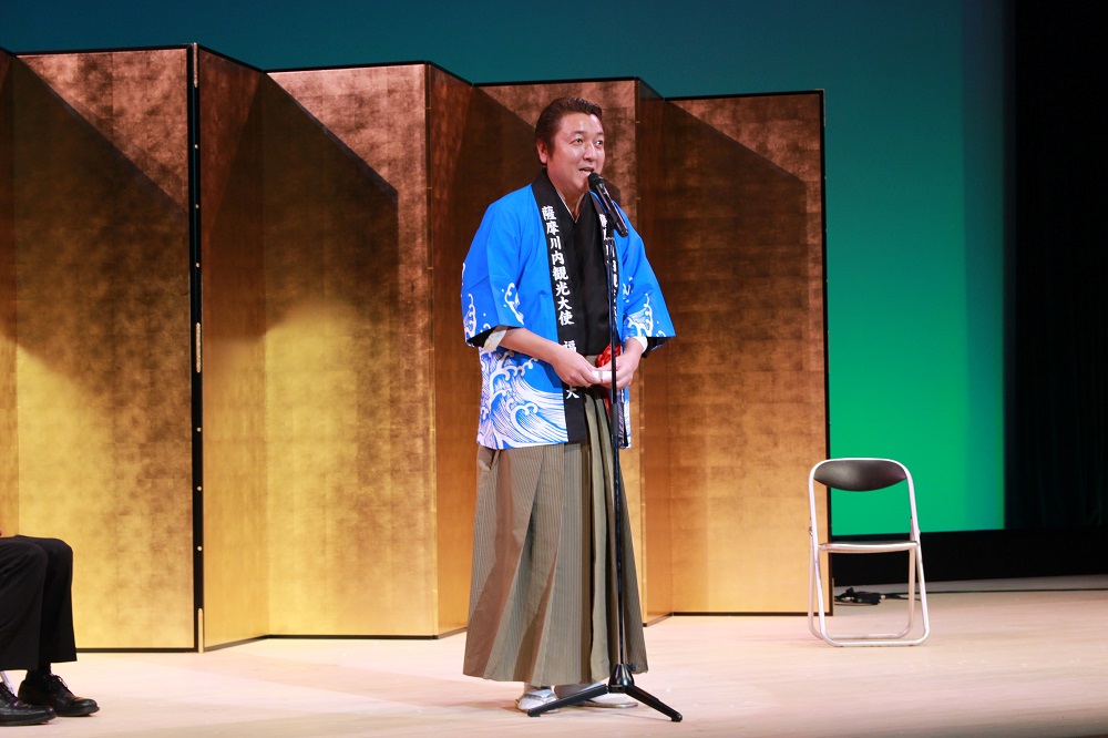 屏風のある壇上で薩摩川内観光大使の法被を着て挨拶する男性の写真
