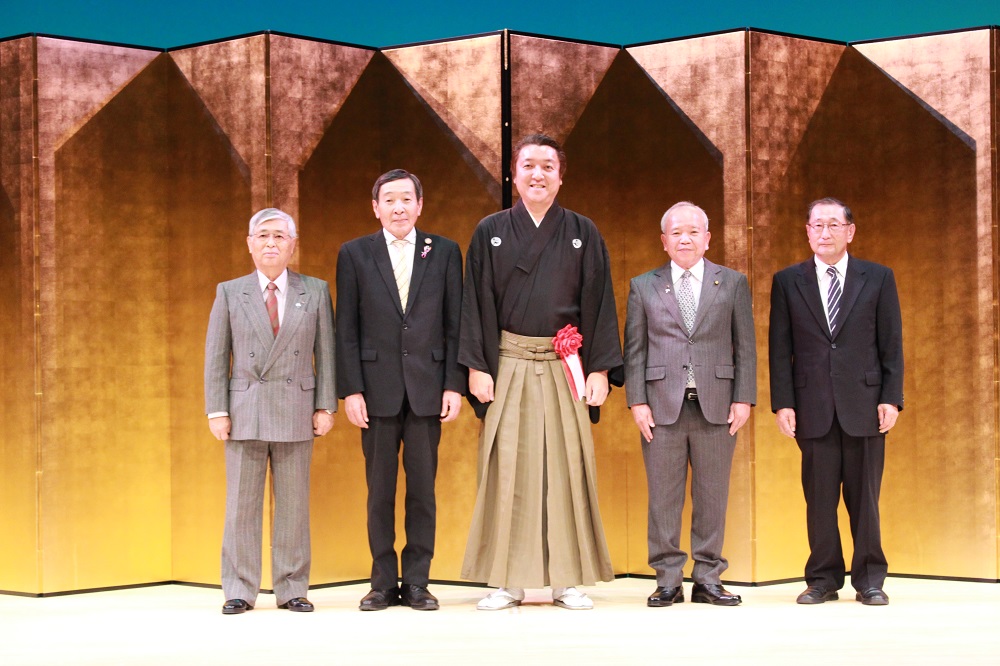 金色の屏風の前で、4人のスーツの中高年の男性が並び、中央に袴姿の男性が立っている写真