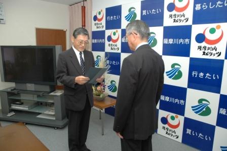 「行きたい」「暮らしたい」「薩摩川内市」と書かれたパネルの前で、スーツ姿の男性がもう一人のスーツ姿の男性に、書面を持ってそれを読み上げている写真