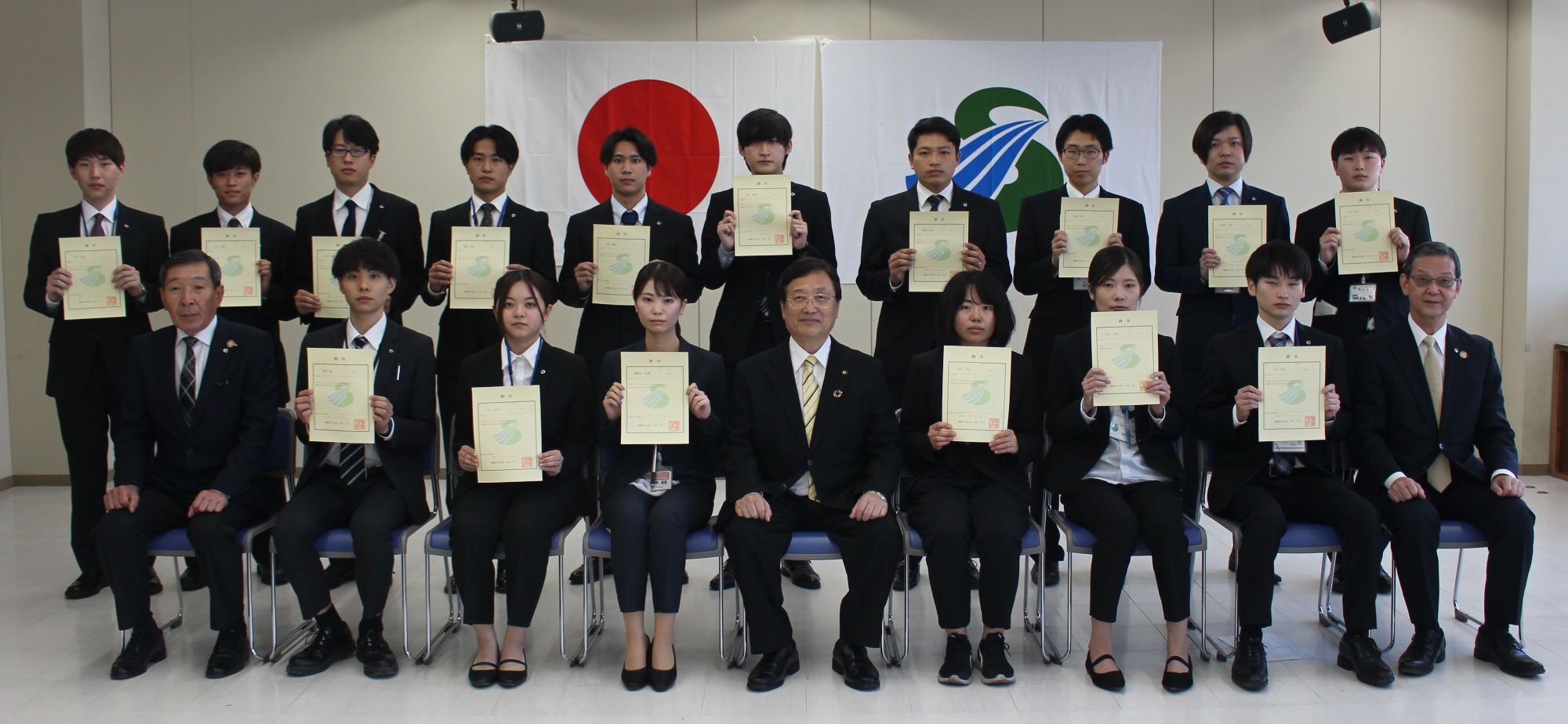 日の丸と薩摩川内市旗が掲げられた室内で、スーツ姿で眼鏡をかけた男性を中心にして、多数の人達が横2列に並び、賞状を両手で持っている記念写真