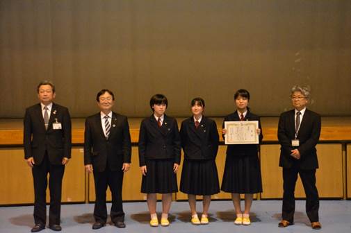 室内でスーツを着た男性3人と女生徒3人が横一列に並んでおり、そのうち女生徒1名が賞状を手に持っている集合写真