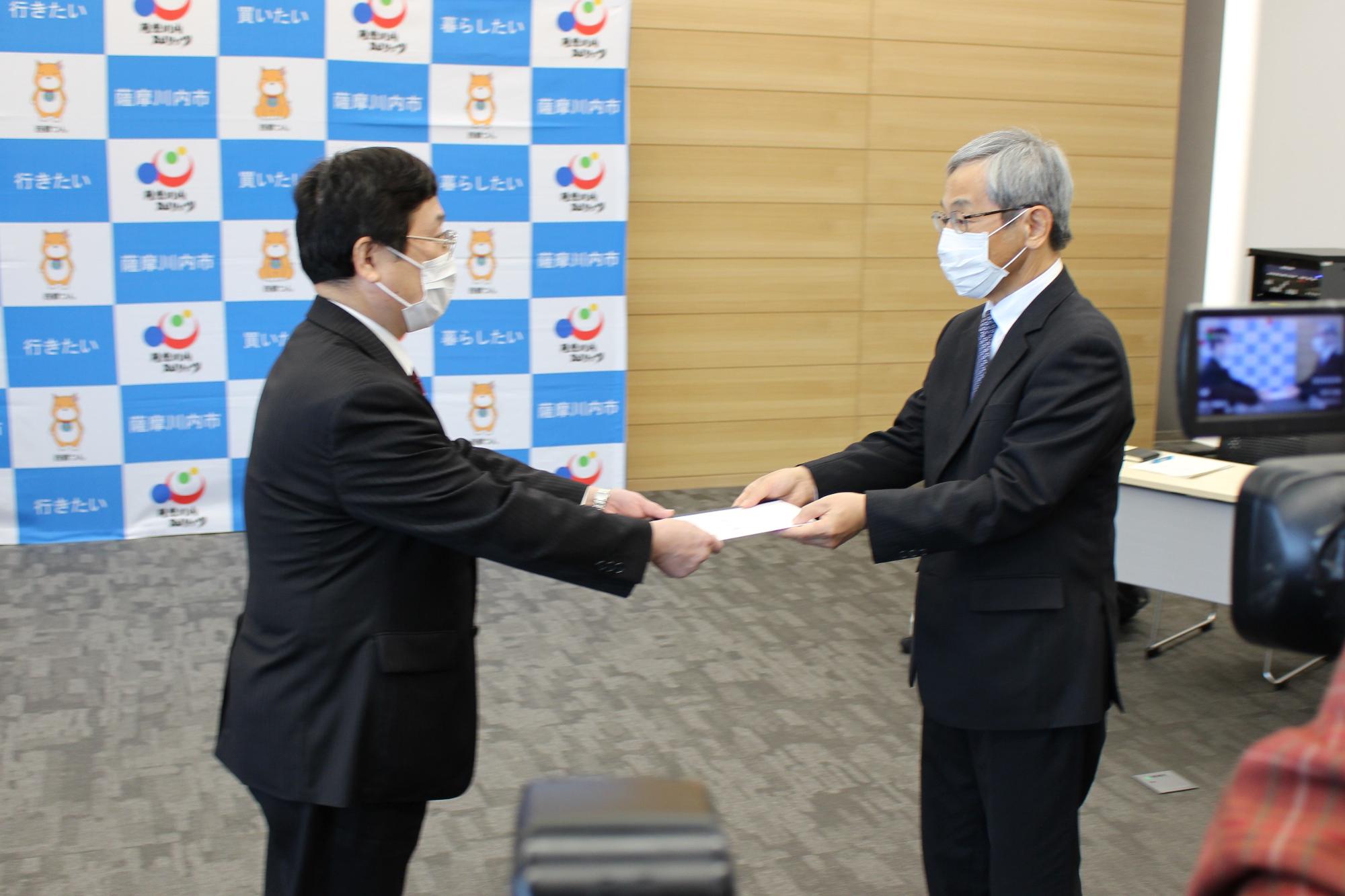 スーツを着た白髪の男性が黒髪の男性に両手で書類を手渡している写真
