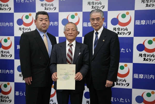 3人の男性がスーツ姿で並び、中央の人物が書類を見えるように掲げている写真